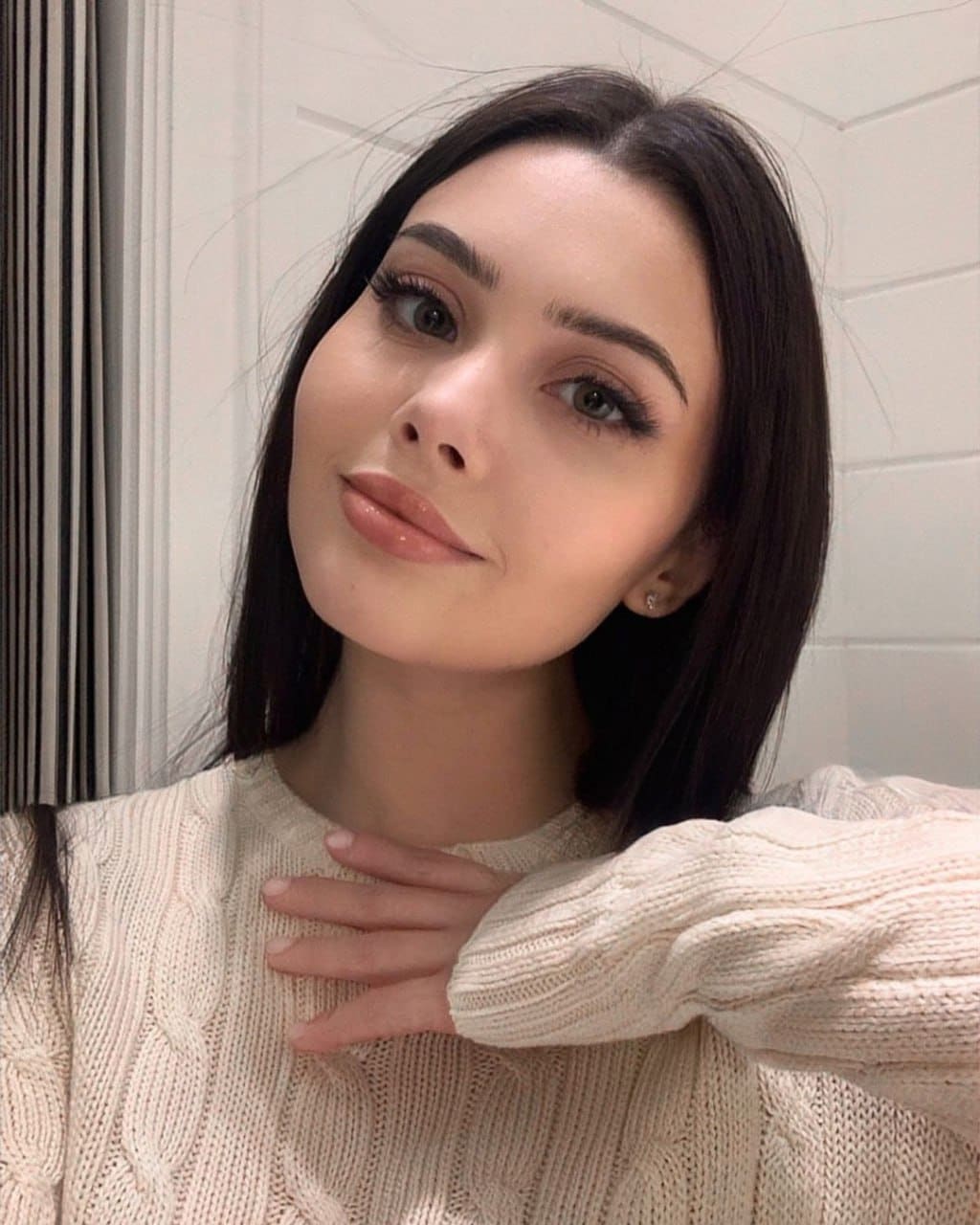 A woman in a cream cable-knit sweater rests a hand on her chest, looking at the camera against a white paneled wall and curtain.