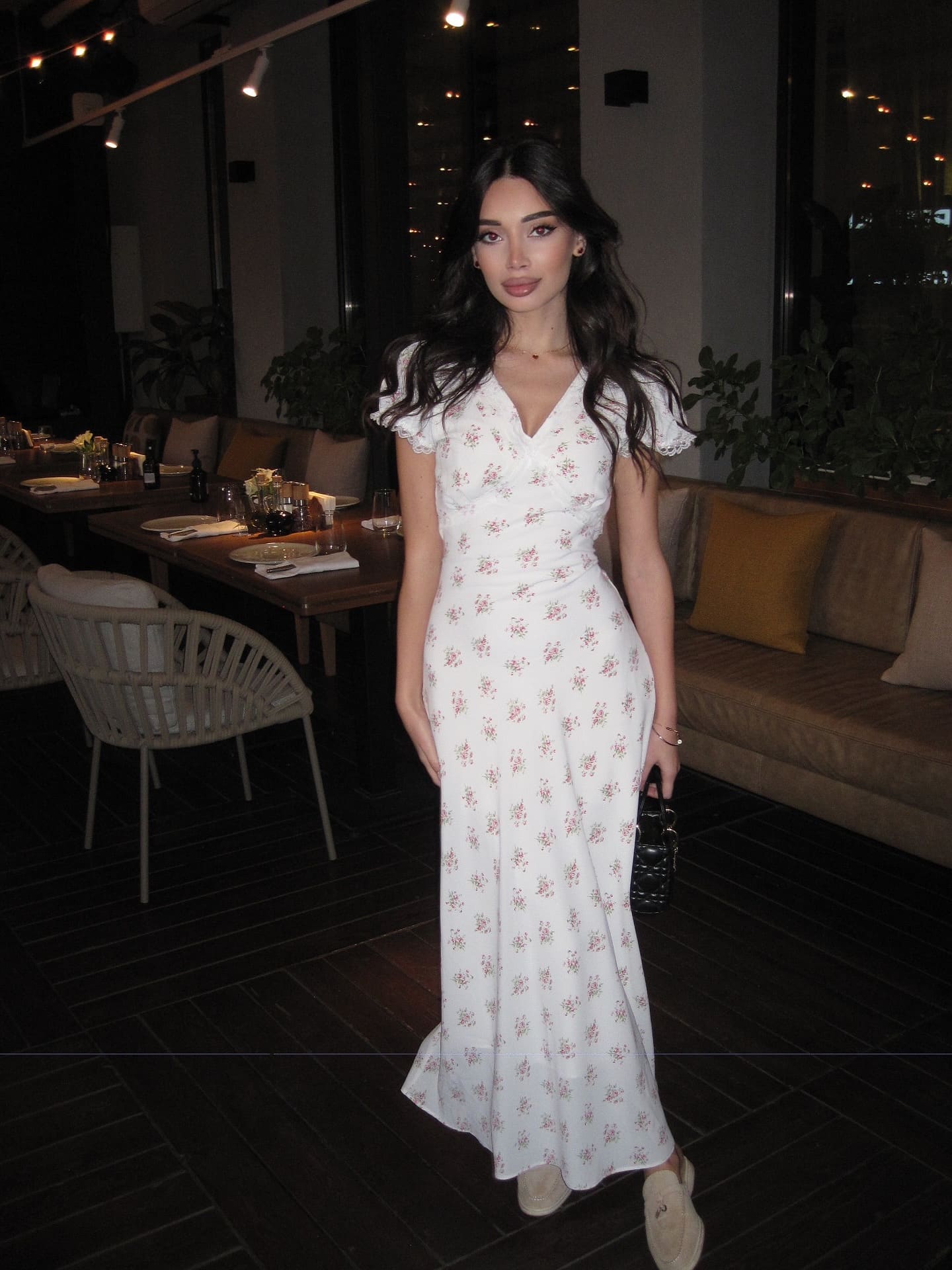 A woman in a white floral midi dress stands in a dim restaurant holding a black handbag, with a dining table behind her.