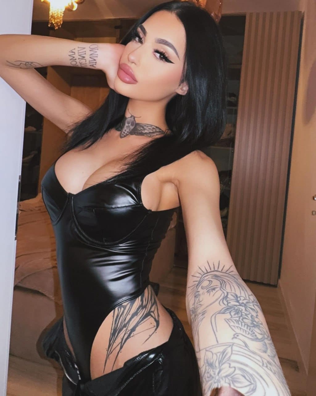 A woman in a black leather bodysuit with cutouts stands indoors near a doorway, with a chandelier visible above and tattoos shown.