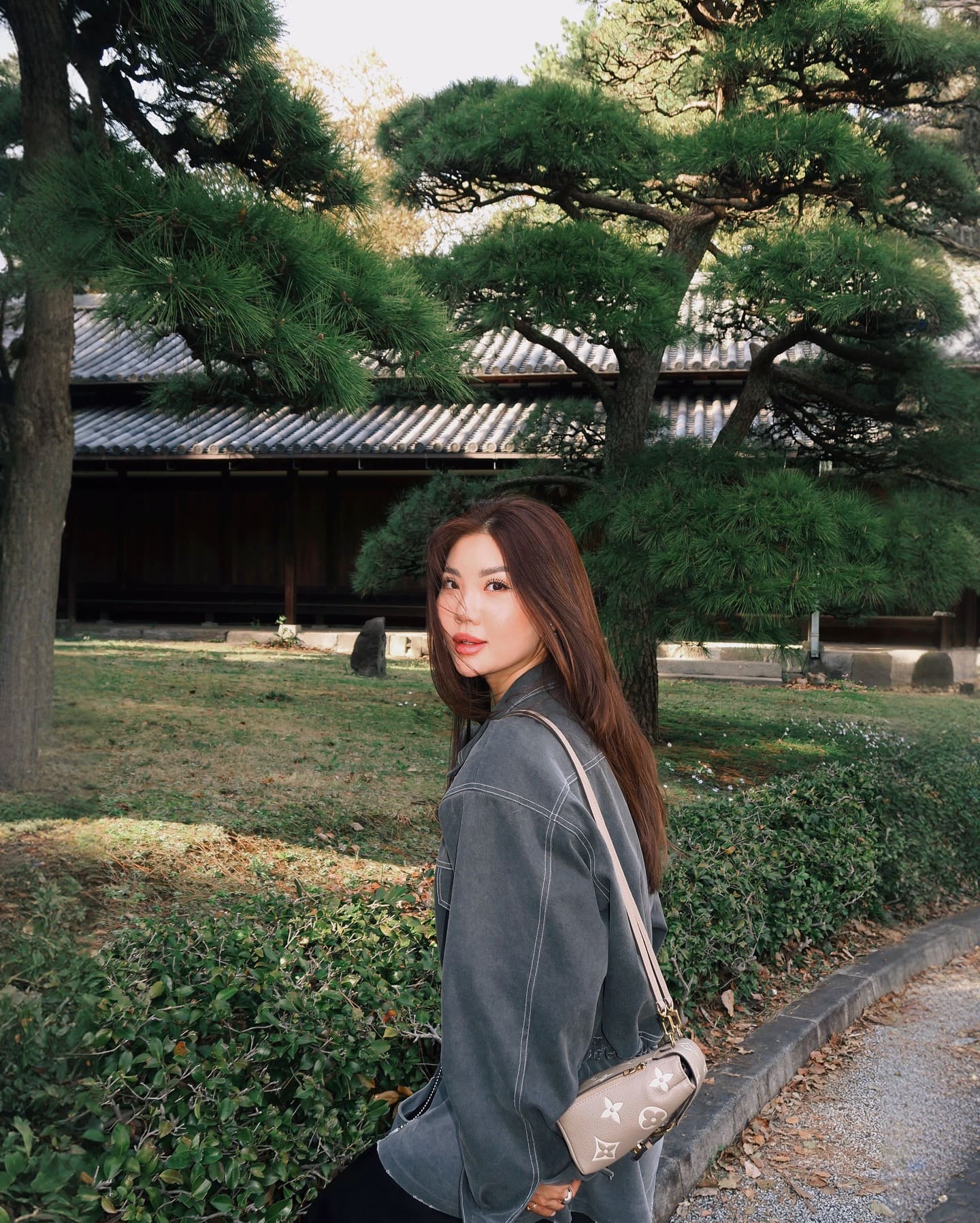 Looking over her shoulder in a garden with pine trees, wearing a grey jacket and small crossbody bag.