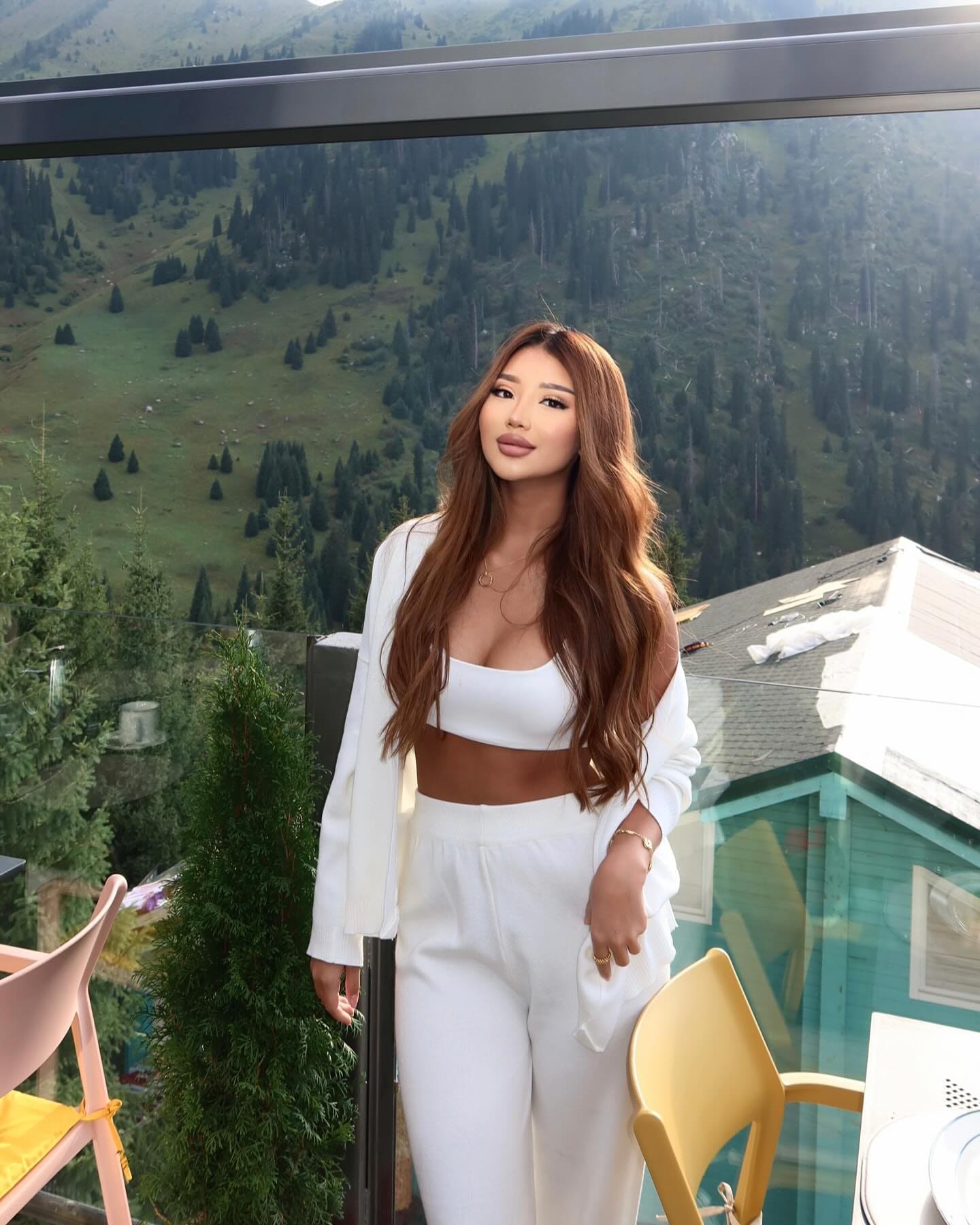 A woman in white trousers and a cardigan stands on a balcony with mountains behind.