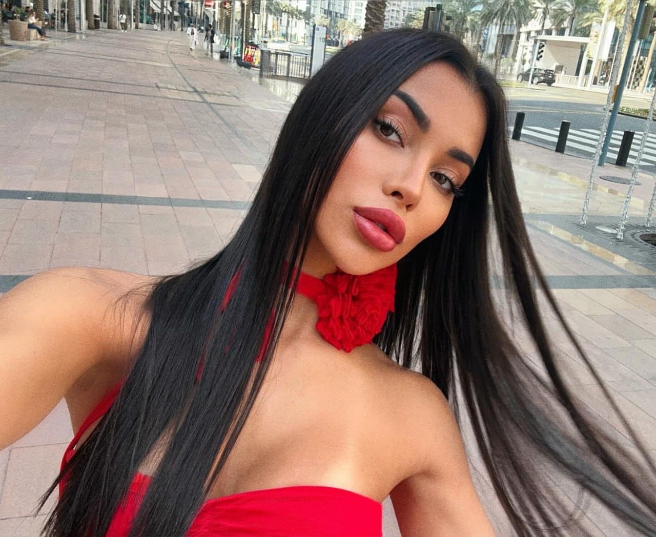 A woman in a red strapless top and ruffled choker poses on a city walkway lined with palm trees and buildings.