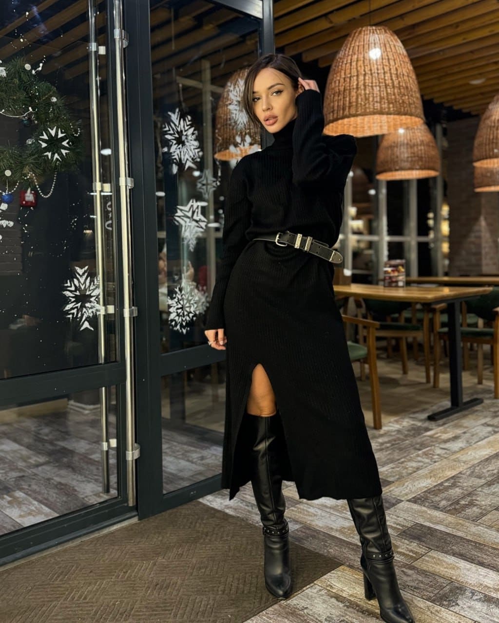 A woman in a black turtleneck dress with a slit and knee-high boots stands by a glass storefront with snowflake decals.