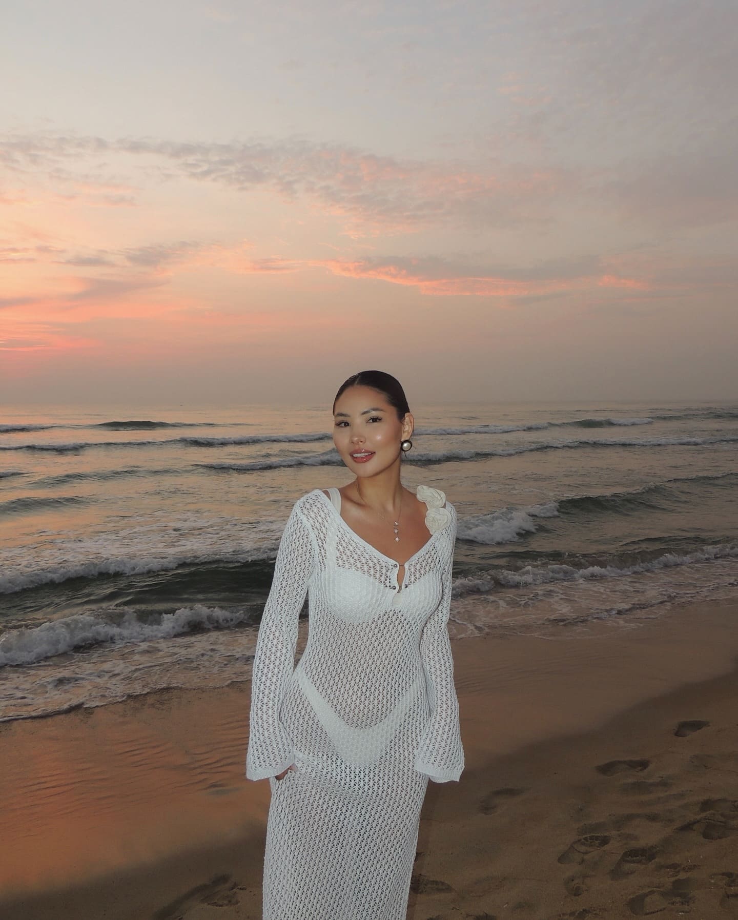 On a beach at sunset, a woman wears a white knit dress with a deep V-neck, ocean and pink sky behind her.
