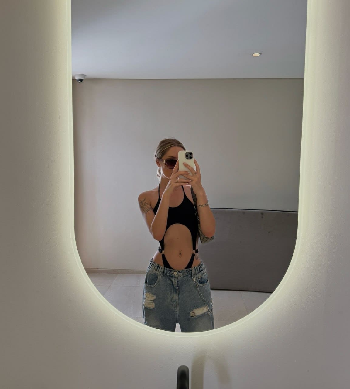 A person with pulled-back hair and sunglasses wears a black cut-out top and jeans for a mirror selfie near a gray bench.