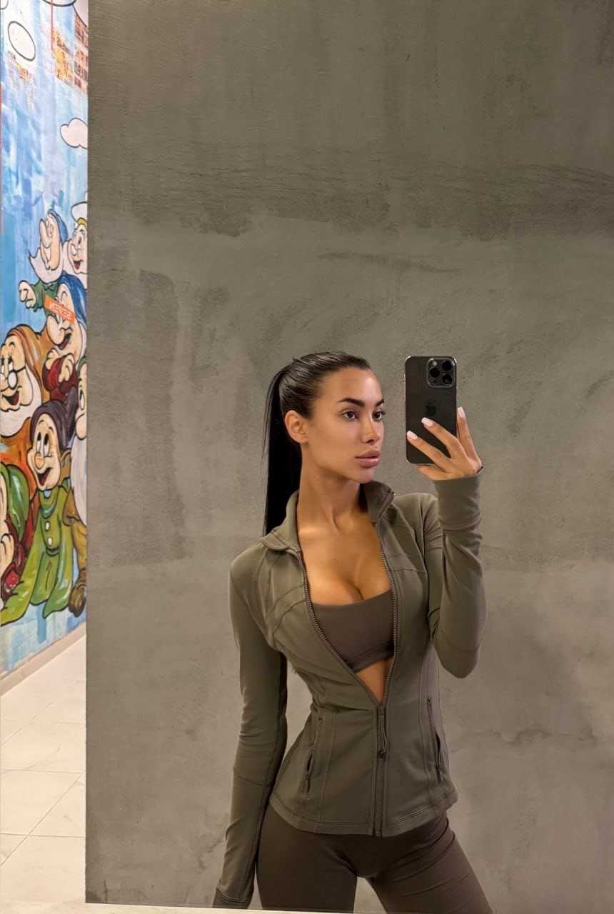 Mirror selfie of a woman in an olive-green athletic jacket and sports bra, standing by a concrete wall with a mural.