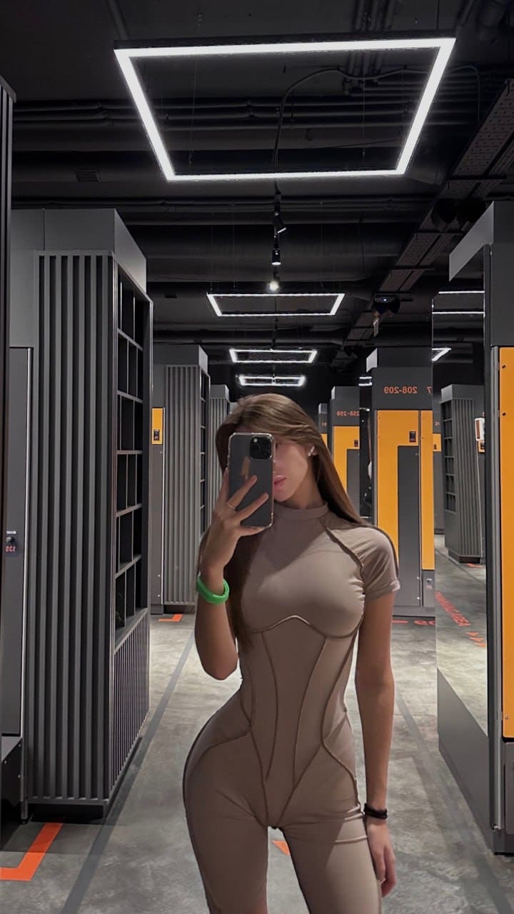 She takes a mirror selfie in a warehouse aisle, wearing a beige bodysuit and holding a smartphone.