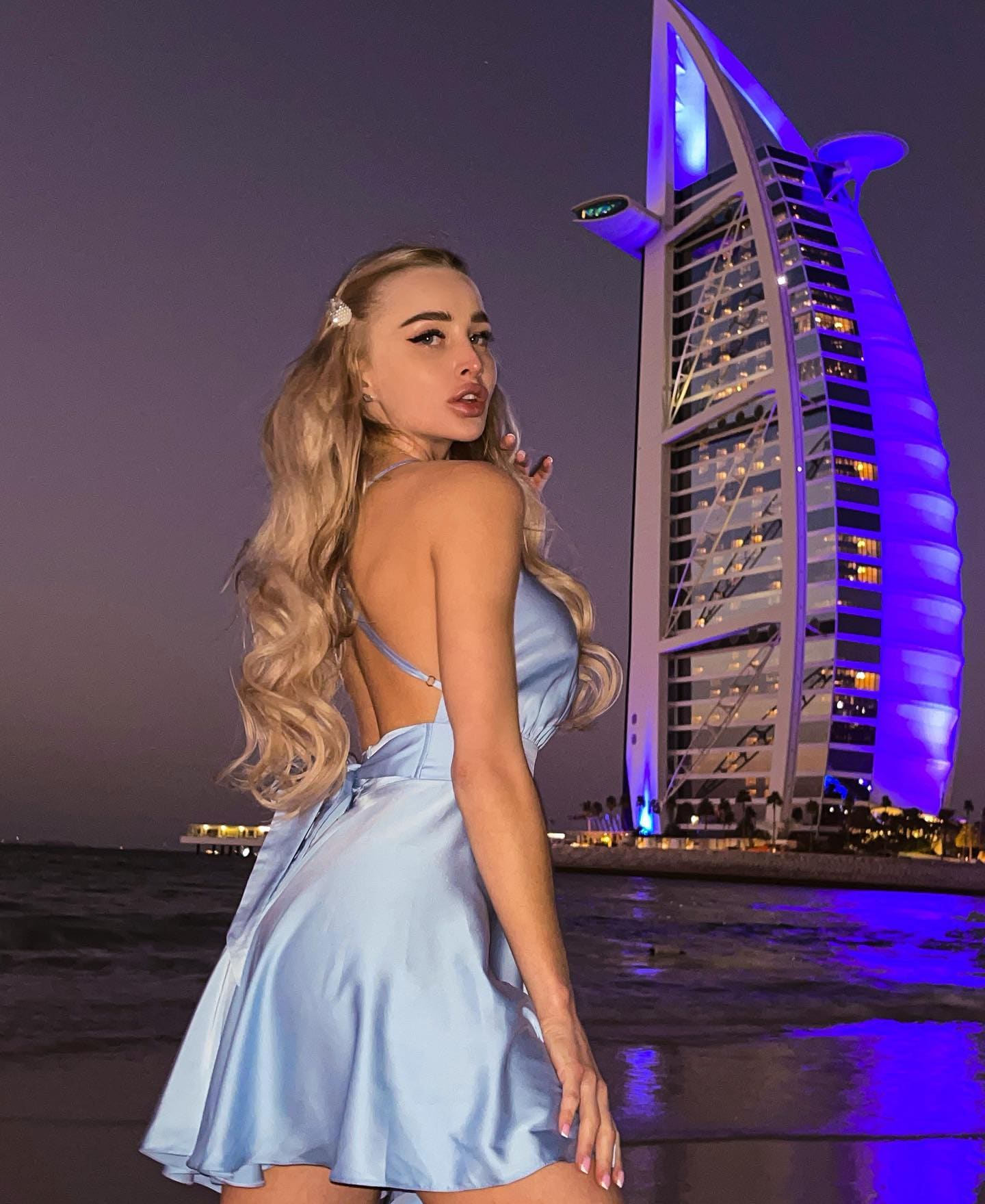 A blonde woman in a light blue satin mini dress with an open back stands on a beach at night, looking over her shoulder with an illuminated building behind.