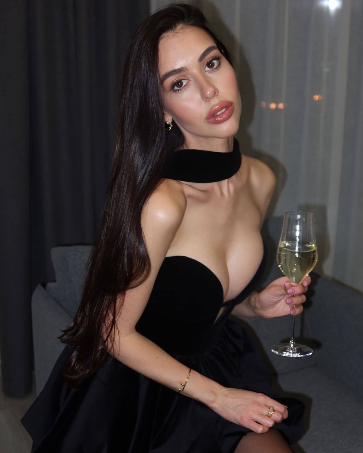 A woman sits on a gray couch in a black strapless dress and choker, holding a glass of white wine while looking at the camera.