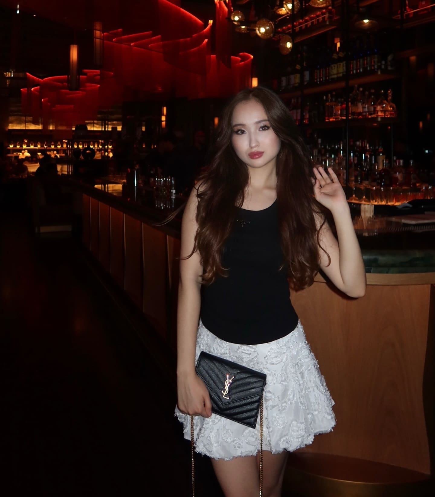 A woman in a black top and white lace skirt holds a clutch in a dimly lit bar, touching her hair with one hand.