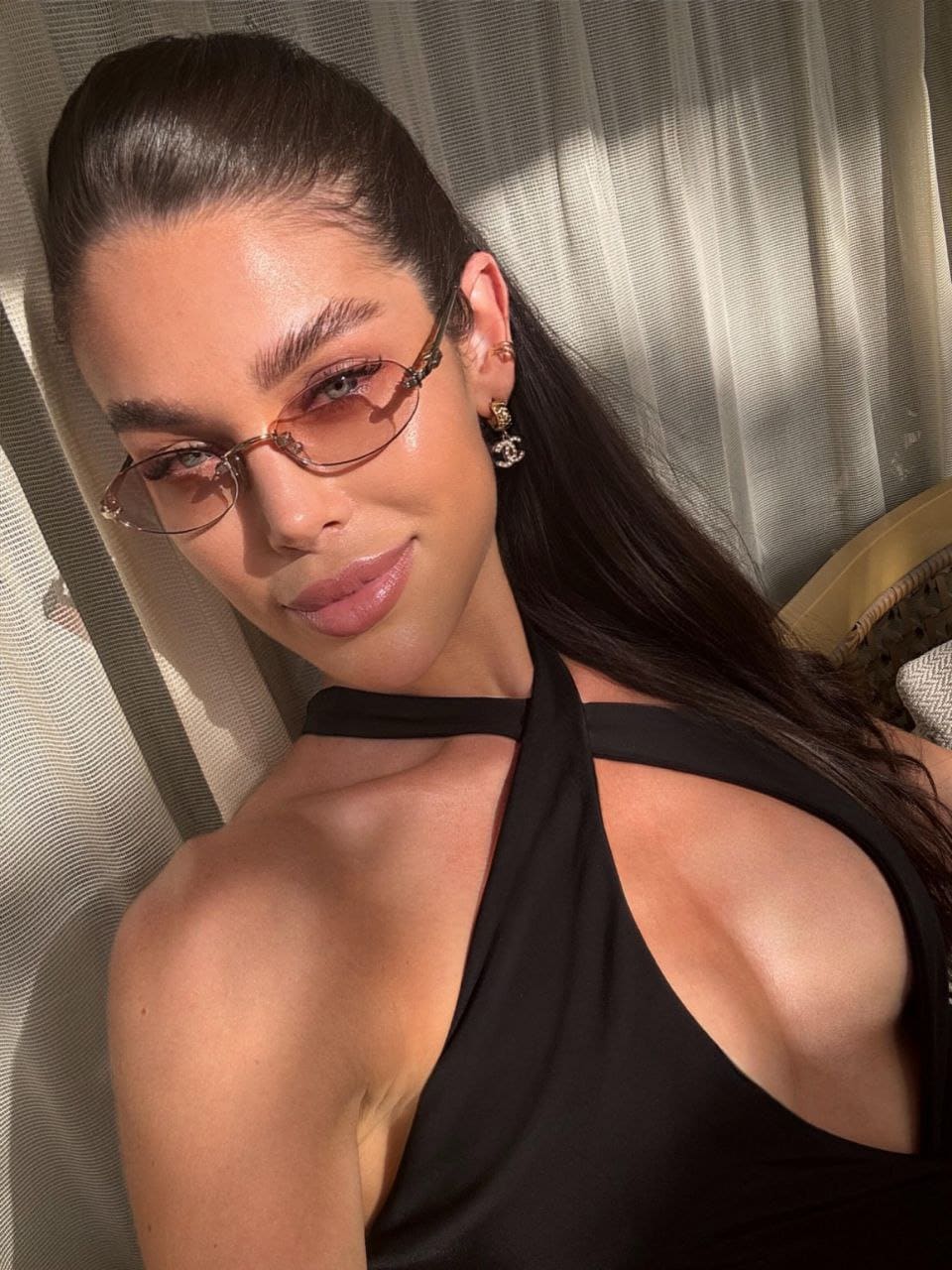 A woman with a ponytail and tinted glasses poses in a black halter top with a shoulder cutout, standing in front of a sheer curtain.