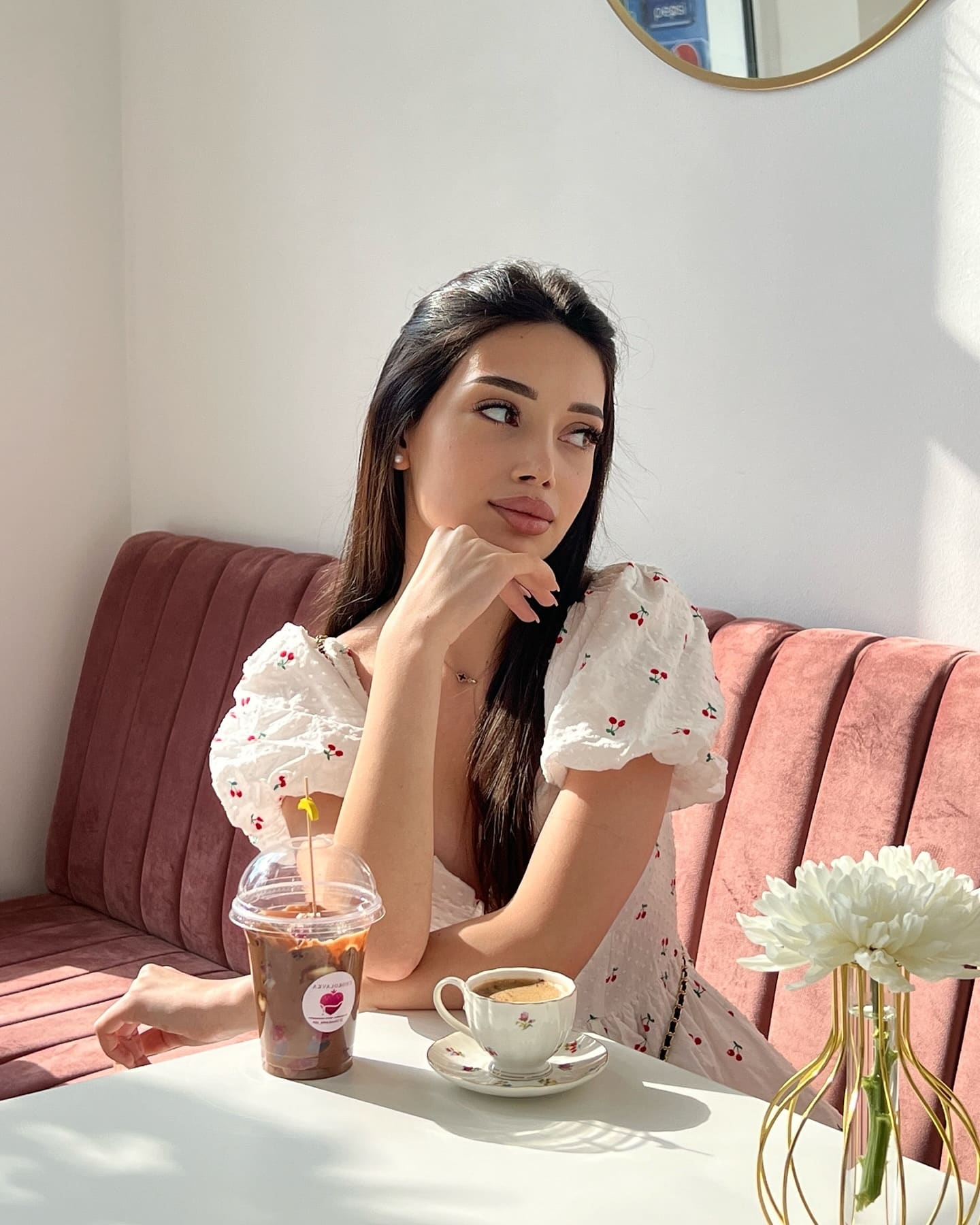 Adelina sits at a cafe table in a white cherry-print dress, resting her chin on her hand beside iced coffee and white flowers.