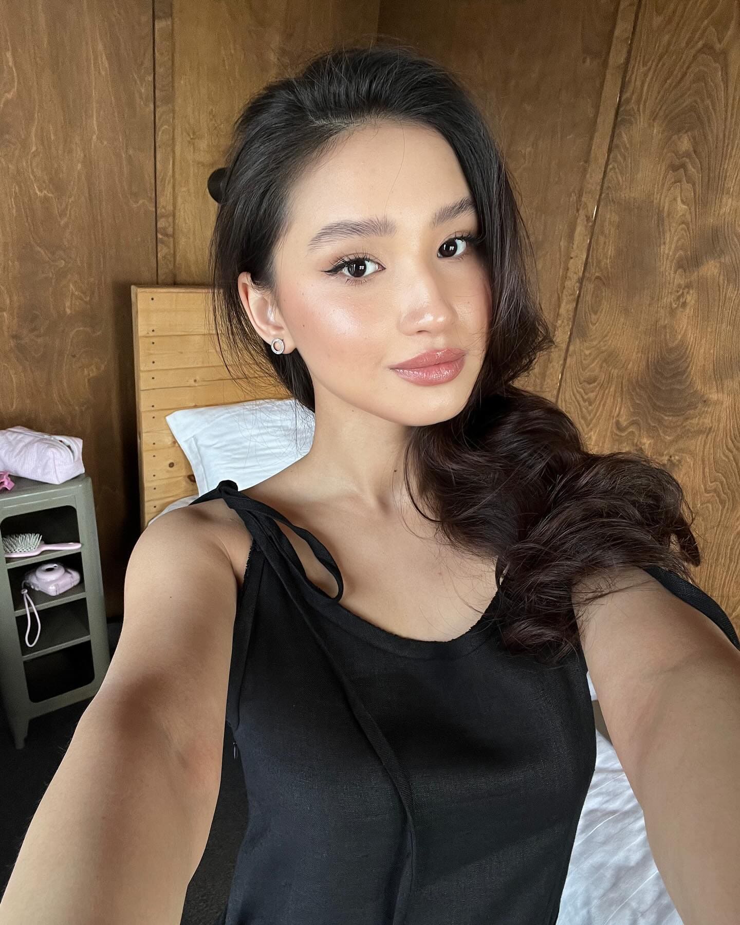 A woman takes a selfie in a wood-paneled room, wearing a black sleeveless top with thin straps and a stud earring.
