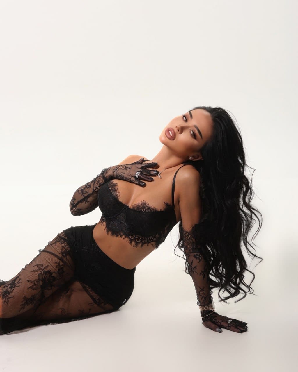 A woman reclines on a white studio floor wearing black lace lingerie and sheer gloves, one hand on her chest.