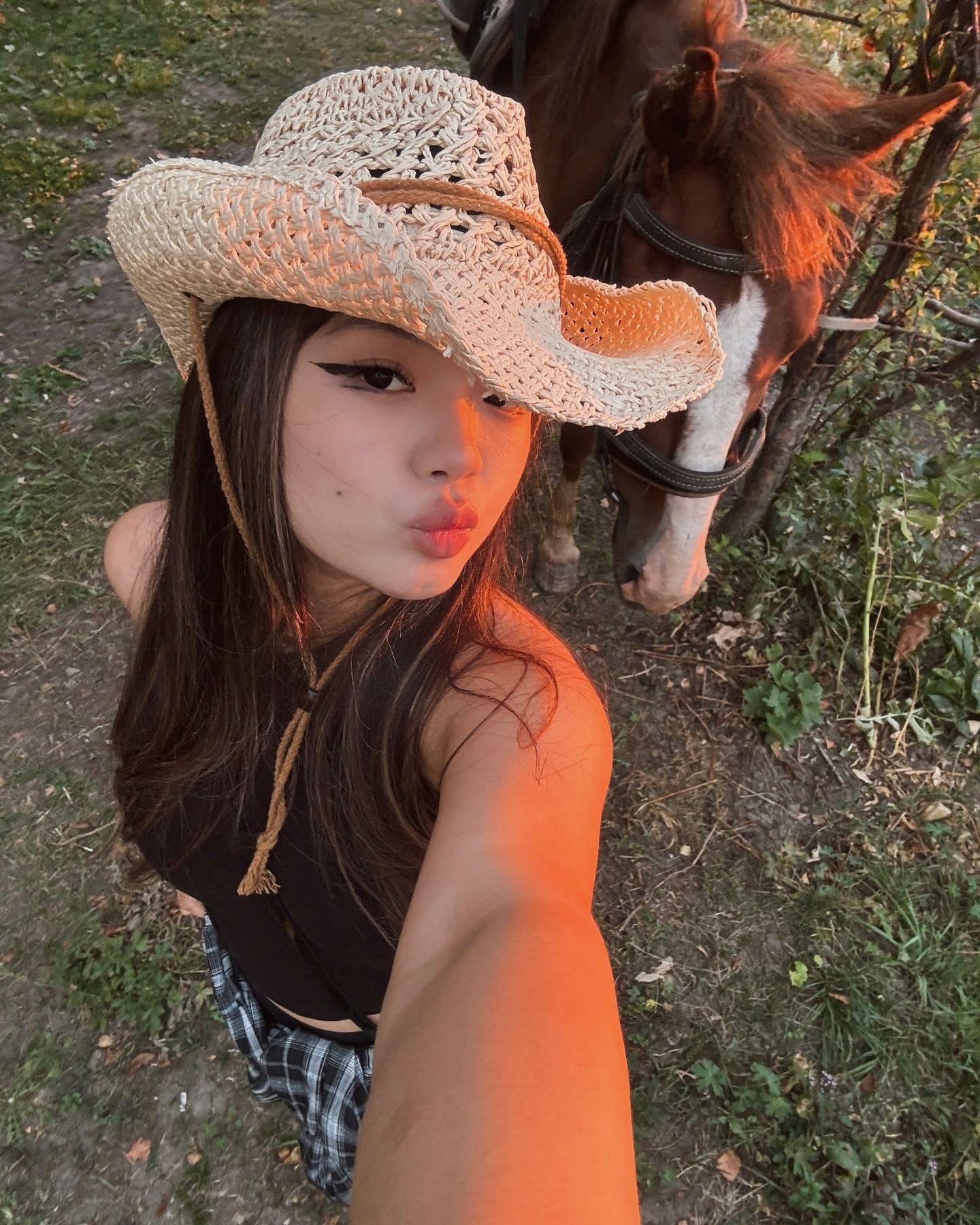 A woman in a straw cowboy hat and black top takes a selfie outdoors, standing beside a brown horse with a white blaze.