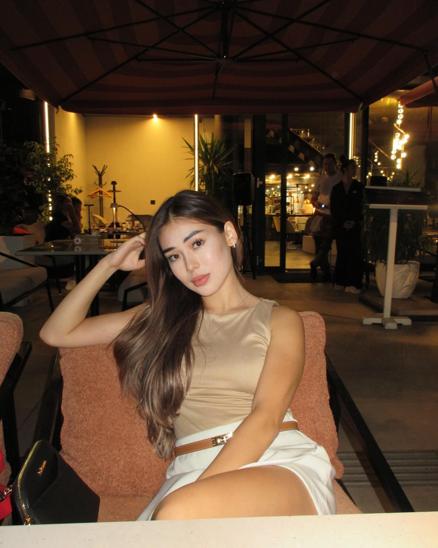 Leila sits on an orange chair outdoors at dusk, wearing a beige top and white skirt, with her hand raised to her long brown hair.
