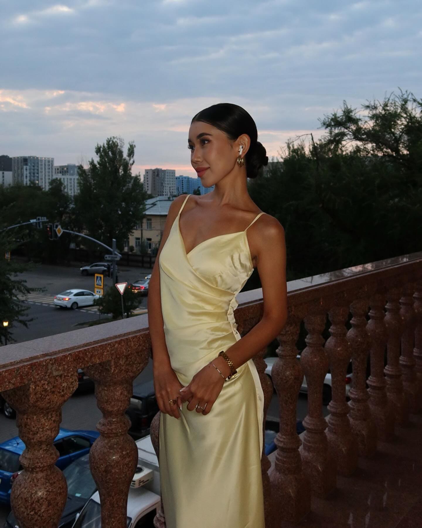 A woman in a pale yellow satin slip dress stands on a stone balcony at dusk, hands clasped, looking away against a city backdrop.