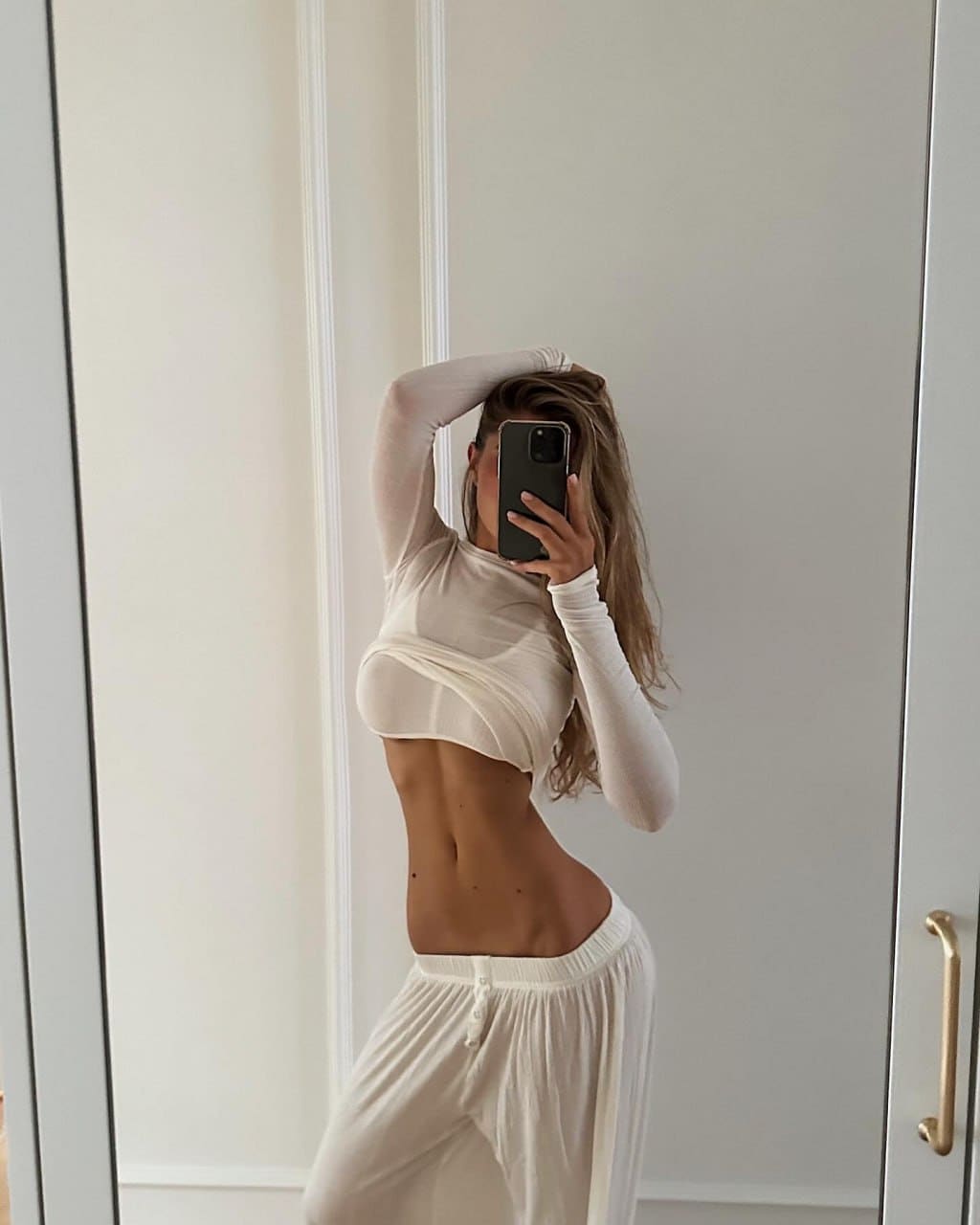 A woman with long wavy brown hair takes a mirror selfie wearing a sheer white long-sleeve crop top and loose white pants, one arm raised behind her head.