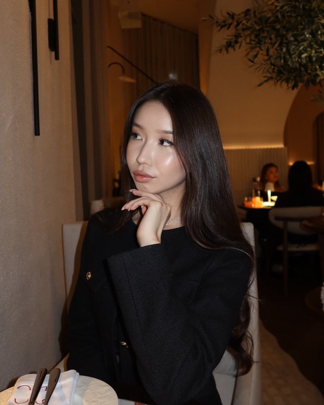 A woman sits at a restaurant table wearing a black textured jacket with gold buttons, resting her chin on her hand while looking aside.
