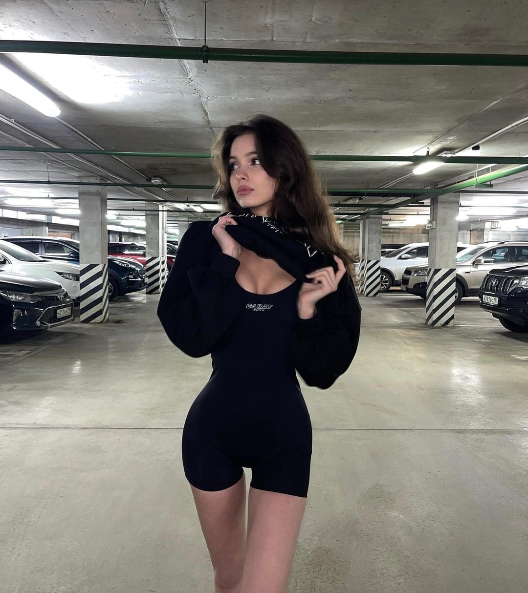 A woman stands in an indoor parking garage wearing a black bodysuit and jacket, with parked cars and concrete pillars visible behind her.