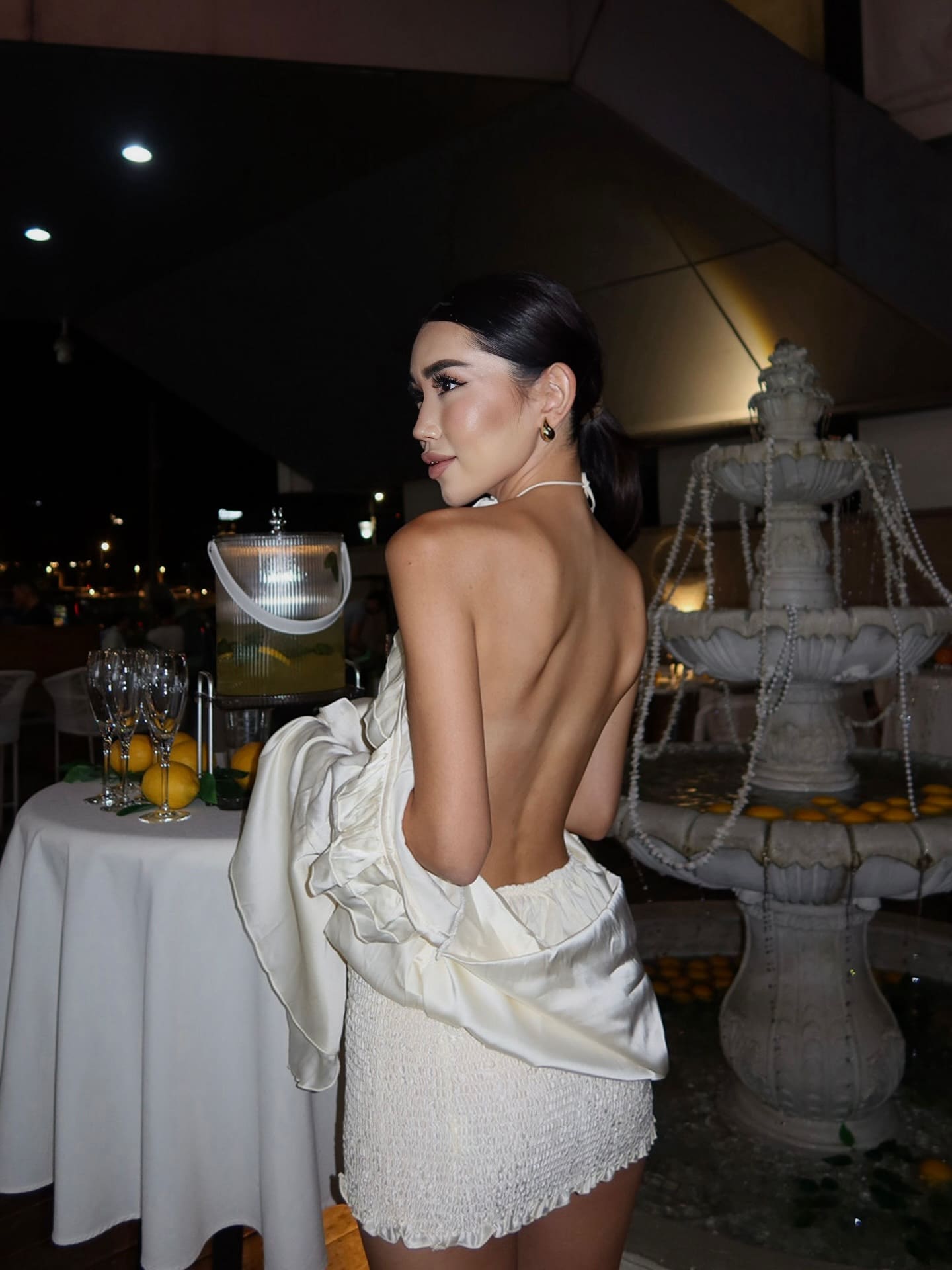 A woman in profile wears a white ruffled off-shoulder top and textured mini skirt, standing near champagne flutes and a fountain.