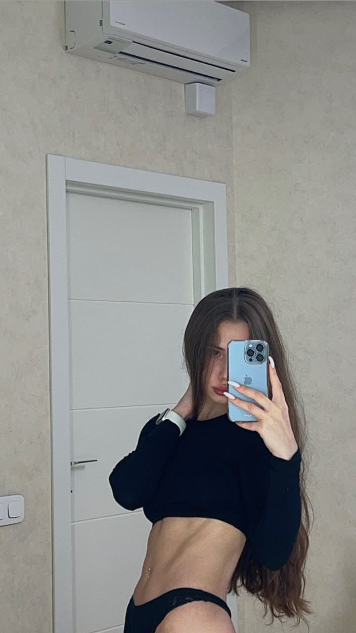 Mirror selfie holding a light blue smartphone, wearing a black long-sleeve crop top and underwear in a room with a white door.