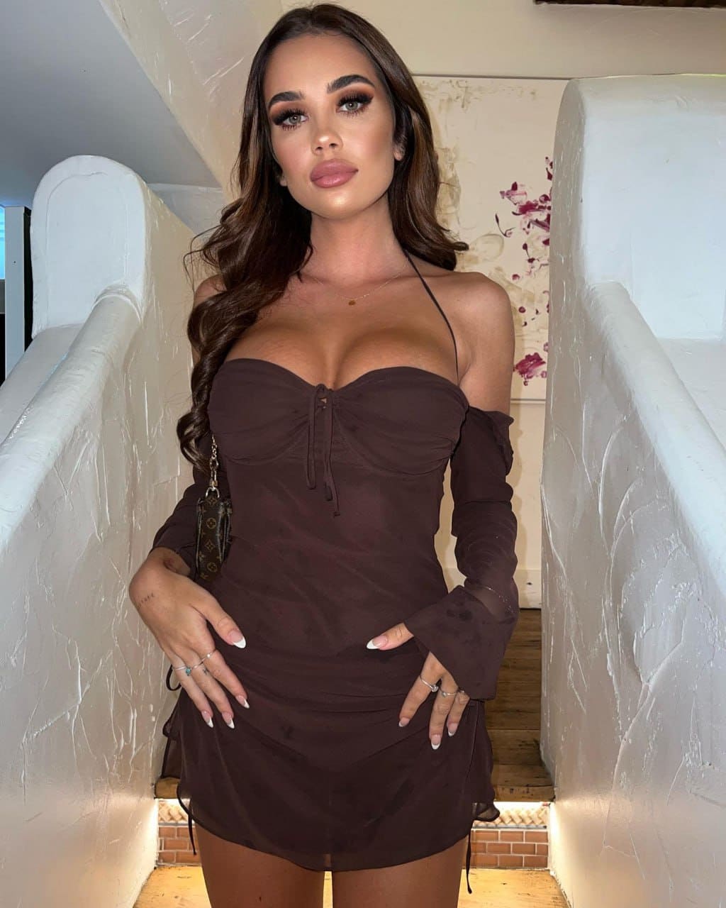Mila stands on a staircase in a dark brown off-the-shoulder mini dress with long sleeves, hands on hips.