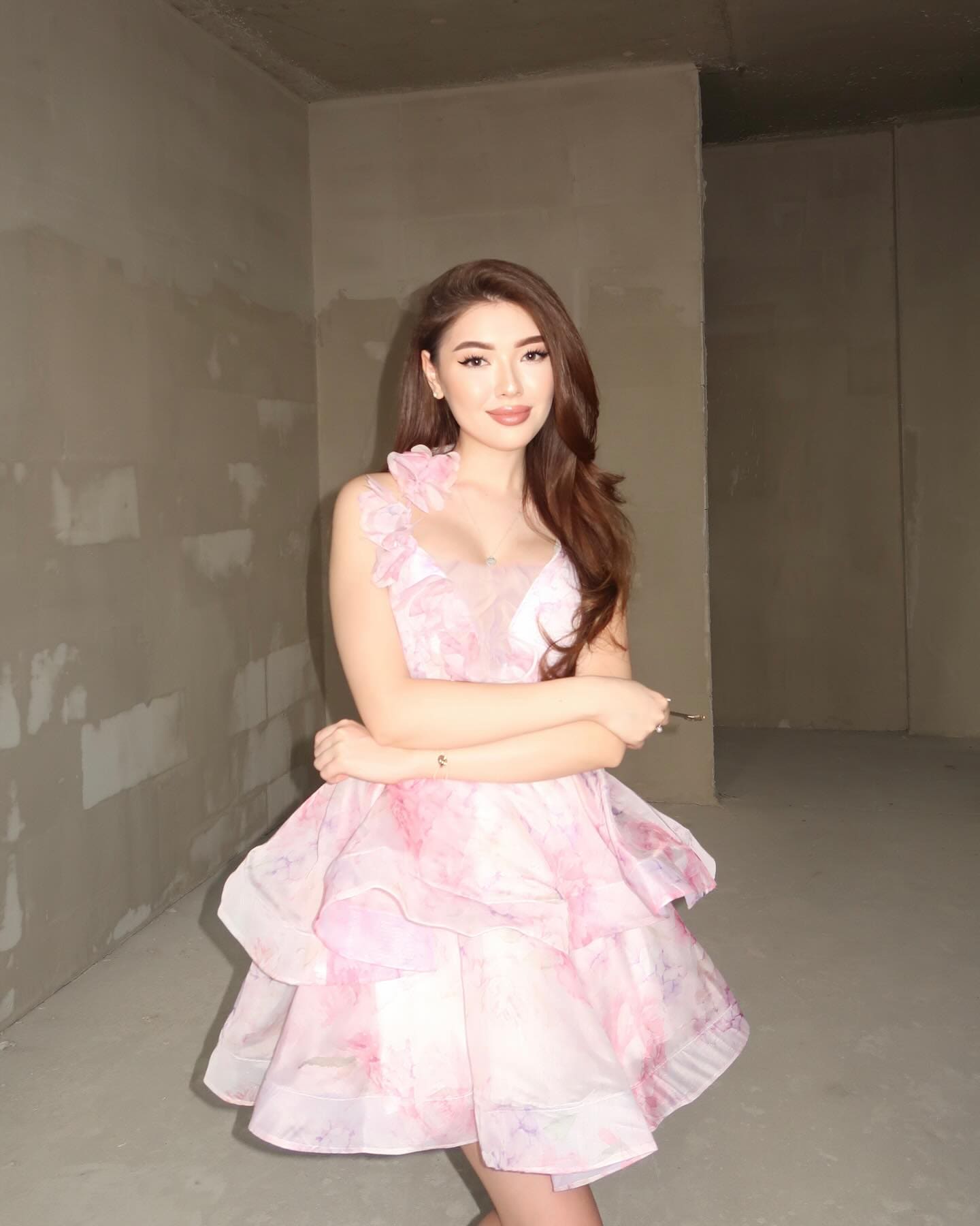 Eliza stands with arms crossed in an unfinished concrete room, wearing a short pink floral dress with ruffled sleeves and a tiered skirt.