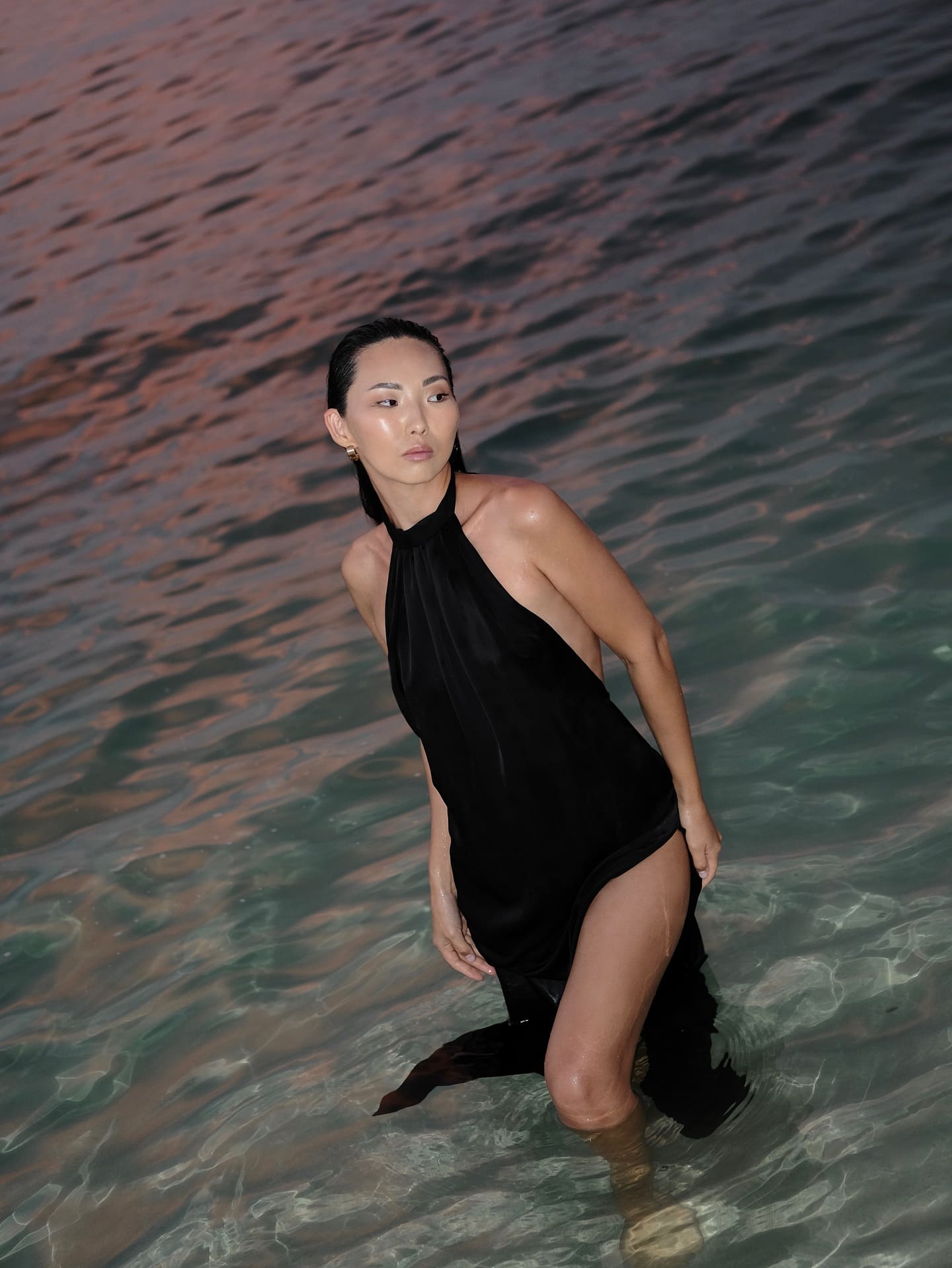 In shallow water, she wears a black halter dress with a high slit, wet hair pulled back, looking toward the camera.