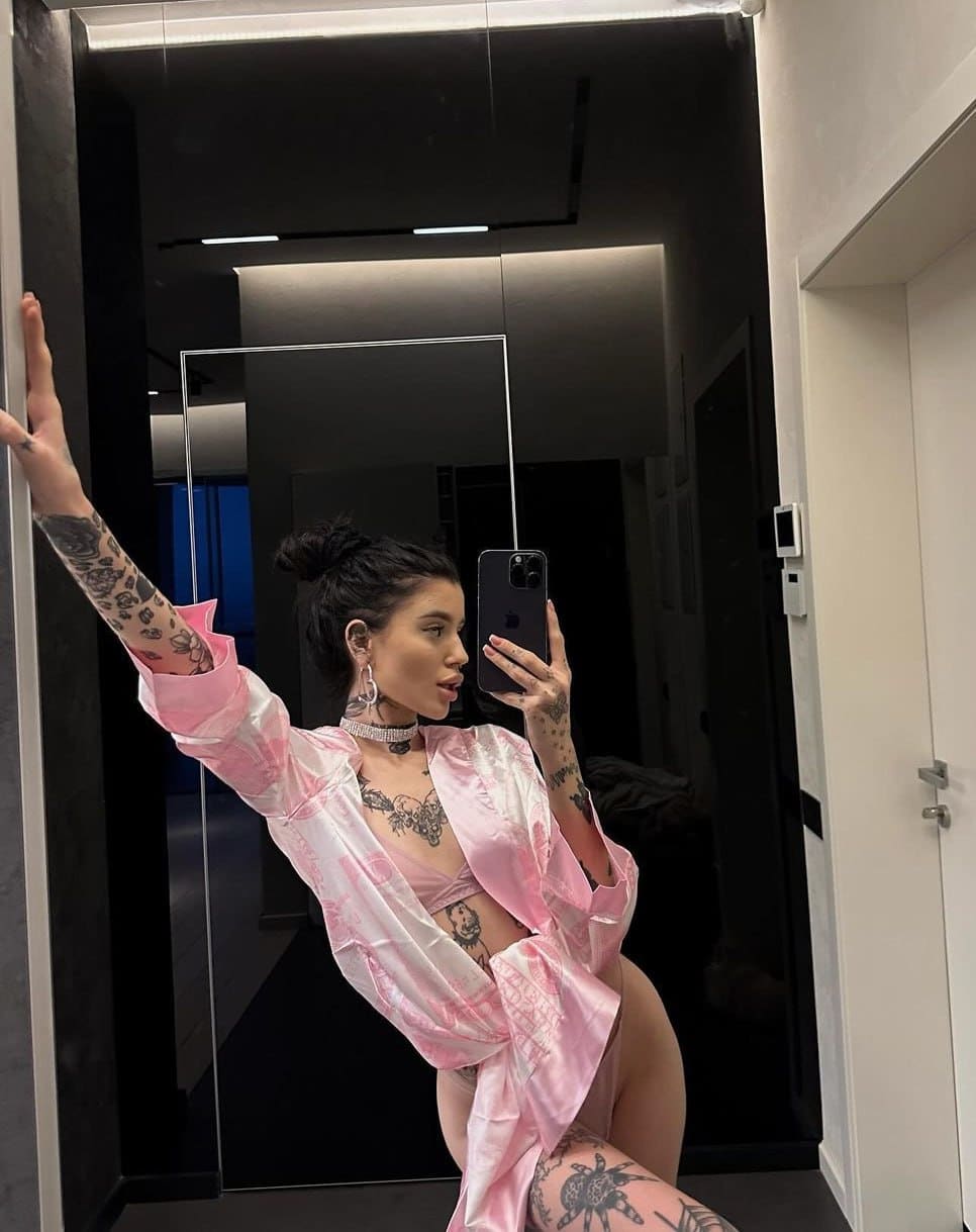A mirror selfie shows a woman with a bun and tattoos in a pink satin robe over a matching bikini top, one arm raised against a dark wall.