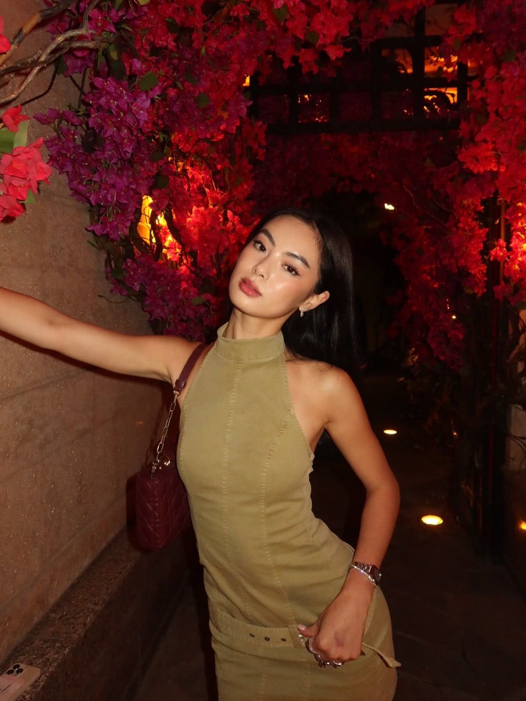 She poses in an olive halter dress with a red bag in a passage lined with pink and red flowers.