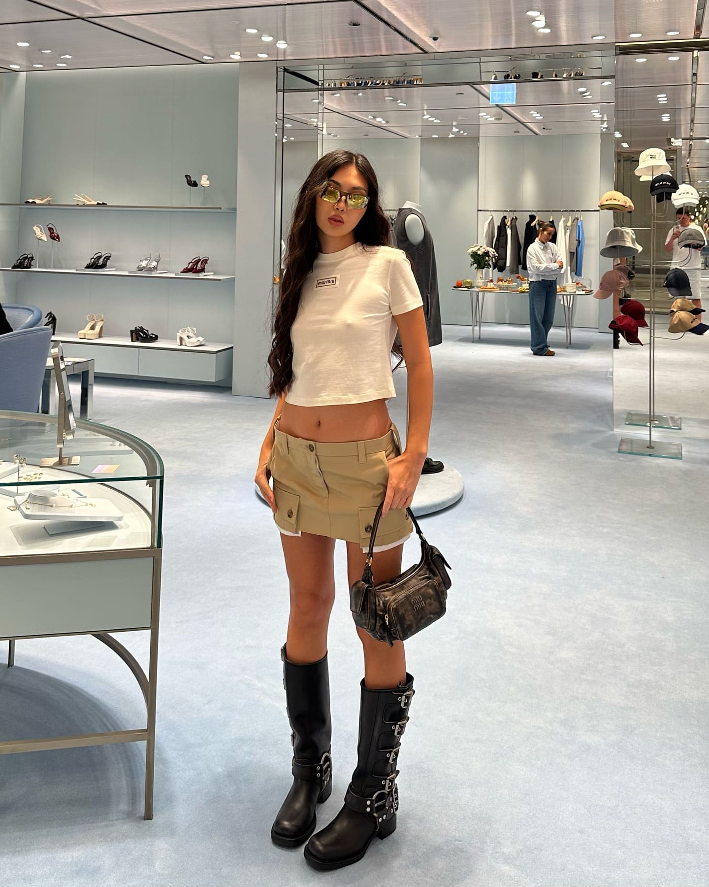 In a store, she wears a white crop top, tan skirt, and knee-high boots, holding a dark bag and wearing yellow sunglasses.