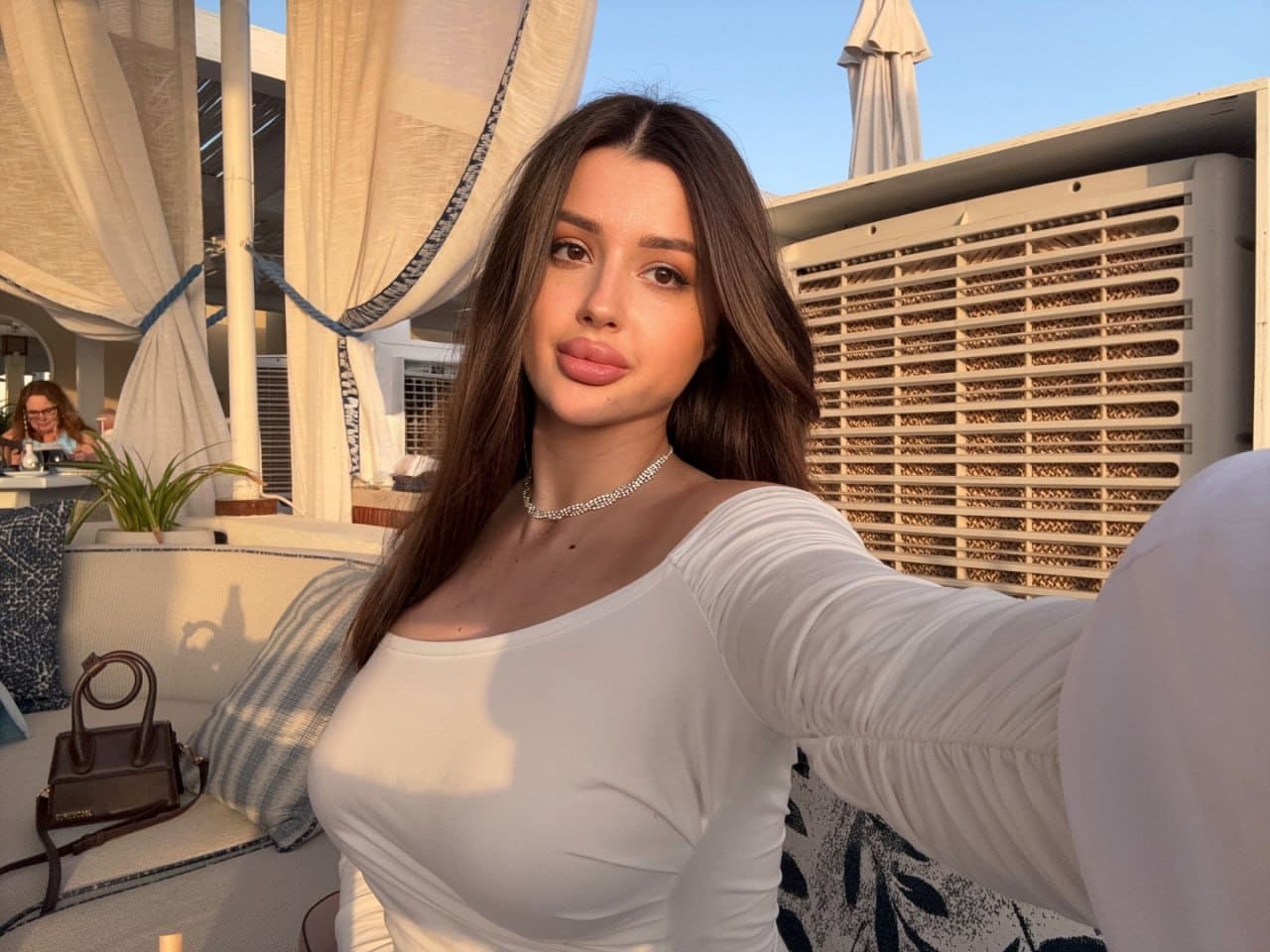 Inna sits on an outdoor lounge with draped curtains, wearing a white long-sleeved top with a wide neckline and a thin necklace.