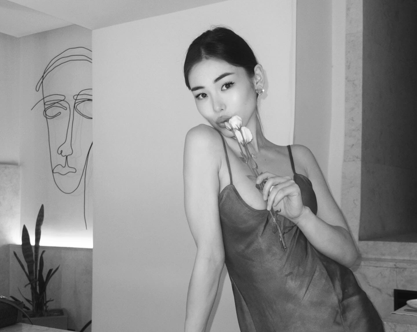 A woman in a slip dress holds a flower to her lips indoors, standing before a wall mural and a potted plant.