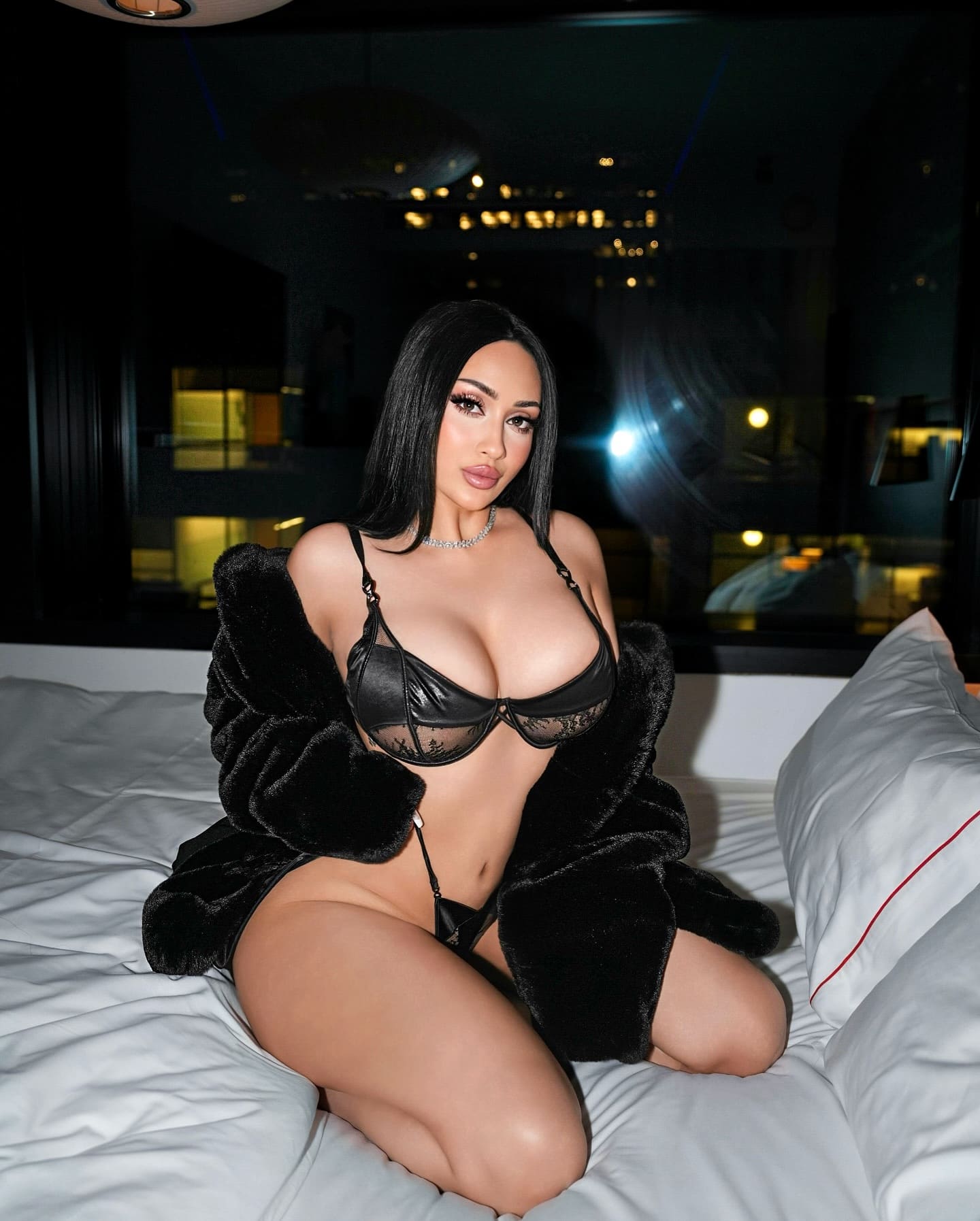 Samfira kneels on a white bed in black lace lingerie and a fur coat, with city lights visible through a dark window behind her.