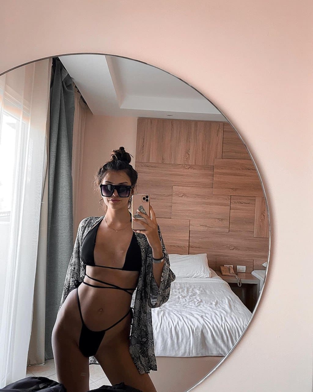 A mirror selfie shows a woman in a black strappy bikini and open patterned robe, wearing sunglasses in a bedroom.