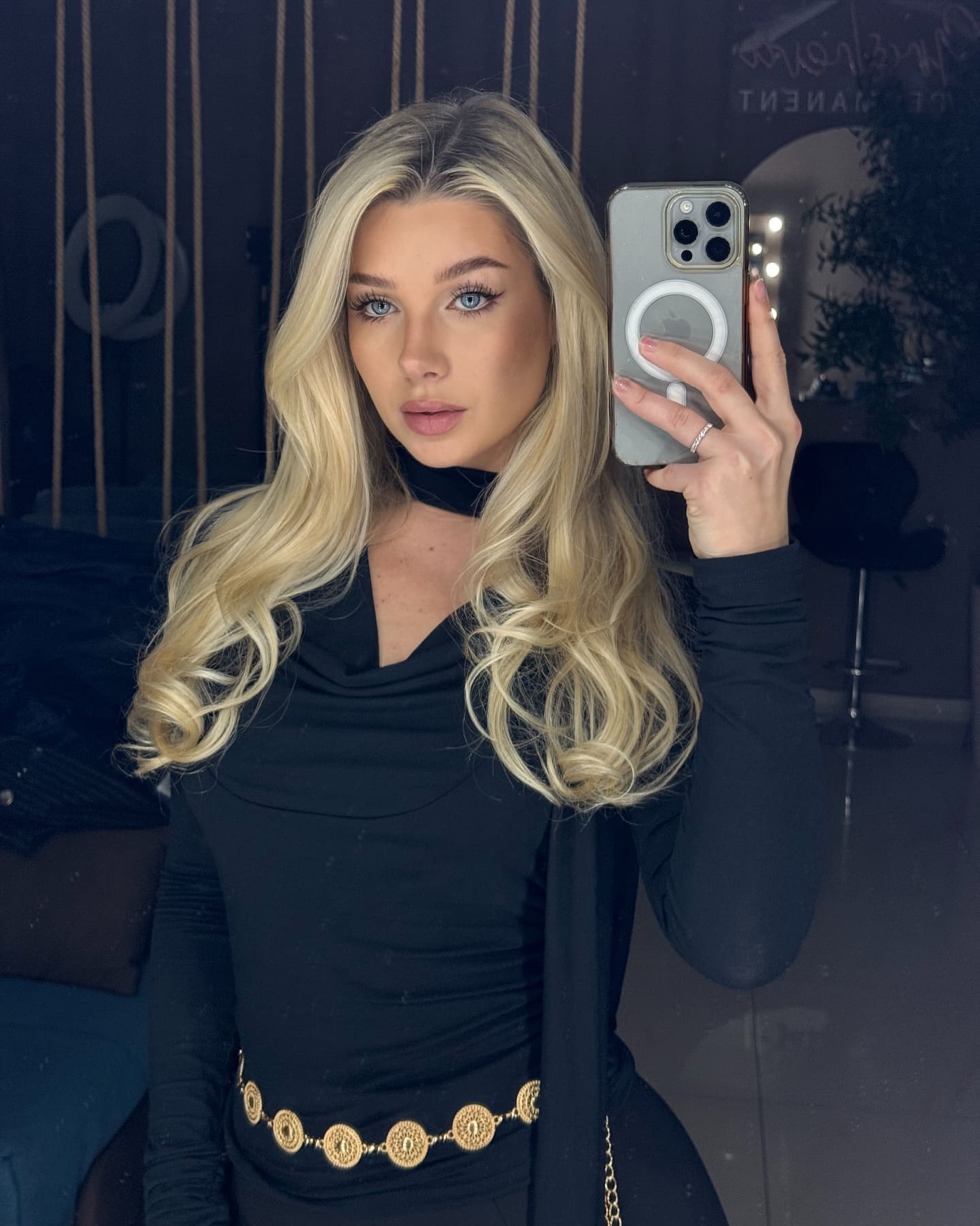 Mirror selfie in a dim room showing a black cowl-neck top with a gold coin belt, with a plant in the background.