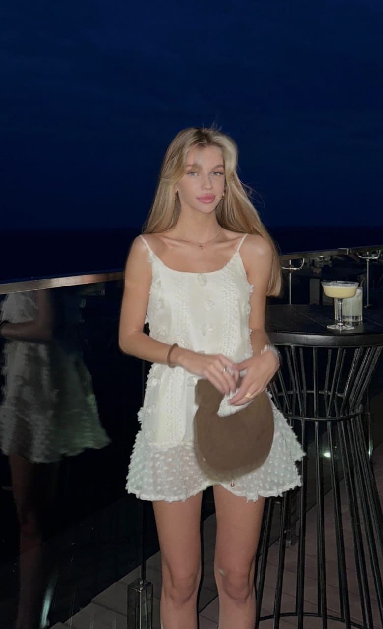 At an outdoor bar at night, she wears a short white dress and holds a brown hat at her waist.