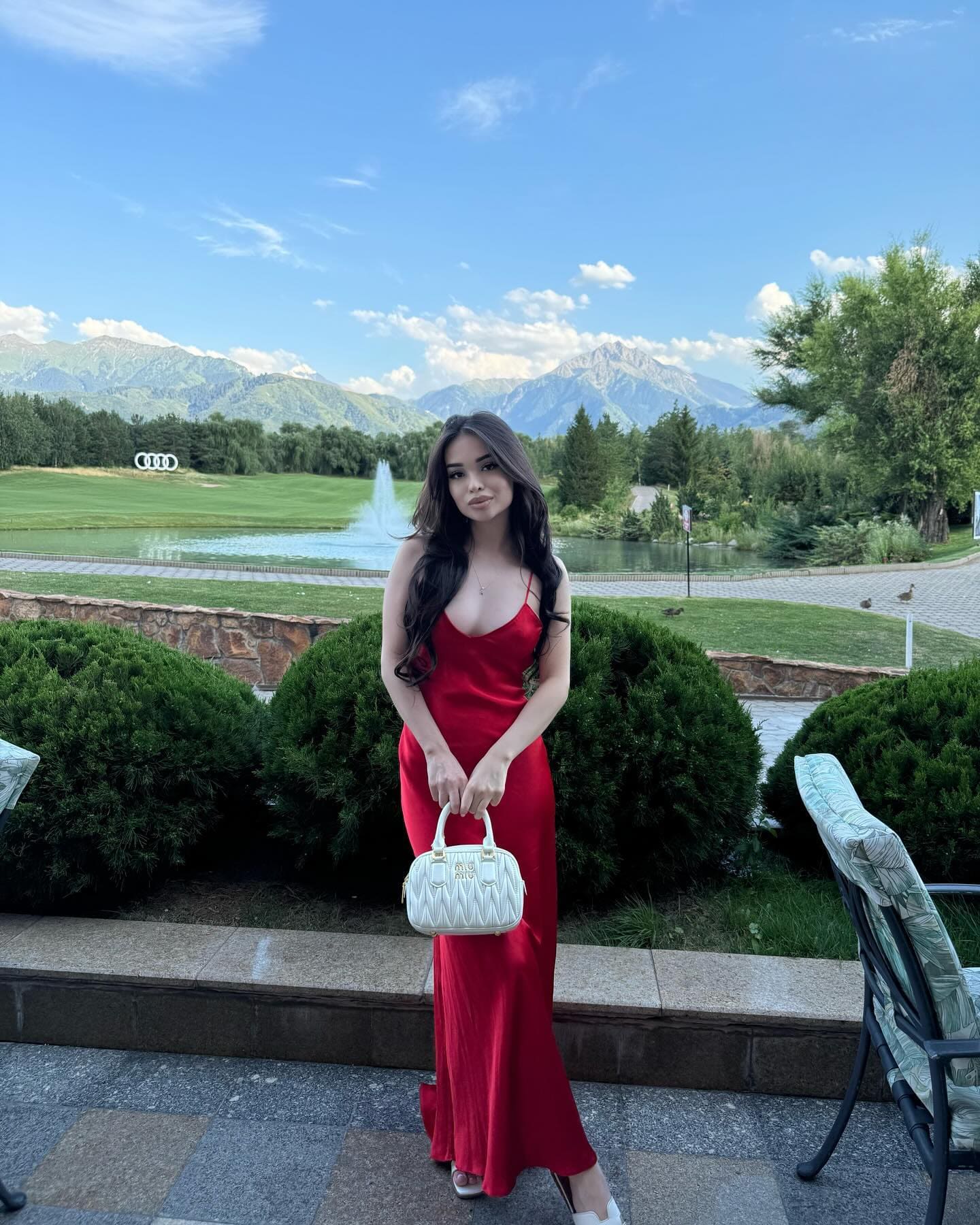 A woman in a red satin maxi dress stands on a stone patio holding a white handbag, with a garden, pond, and mountains behind.