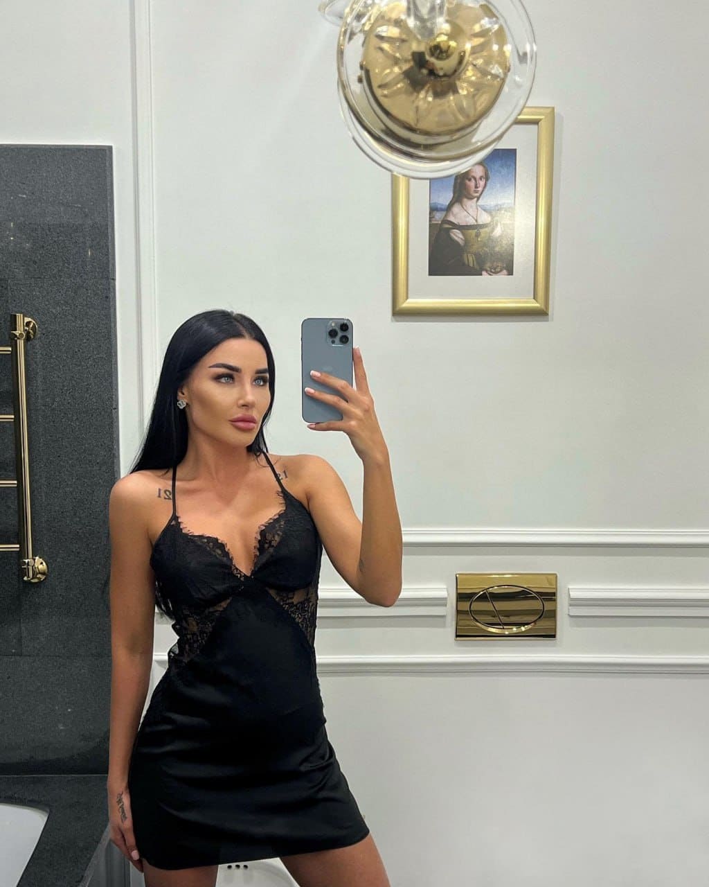 A mirror selfie shows a woman in a short black lace-trimmed slip dress in a bathroom with a white paneled wall.