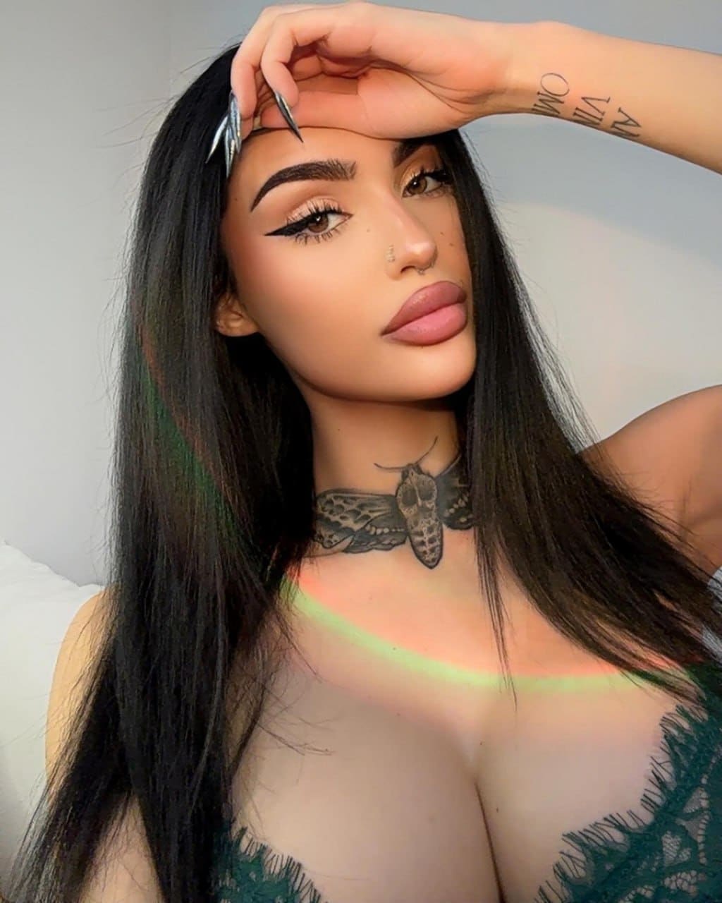 A woman with a neck tattoo wears a dark green lace top, posing with a hand to her forehead against a plain light background.
