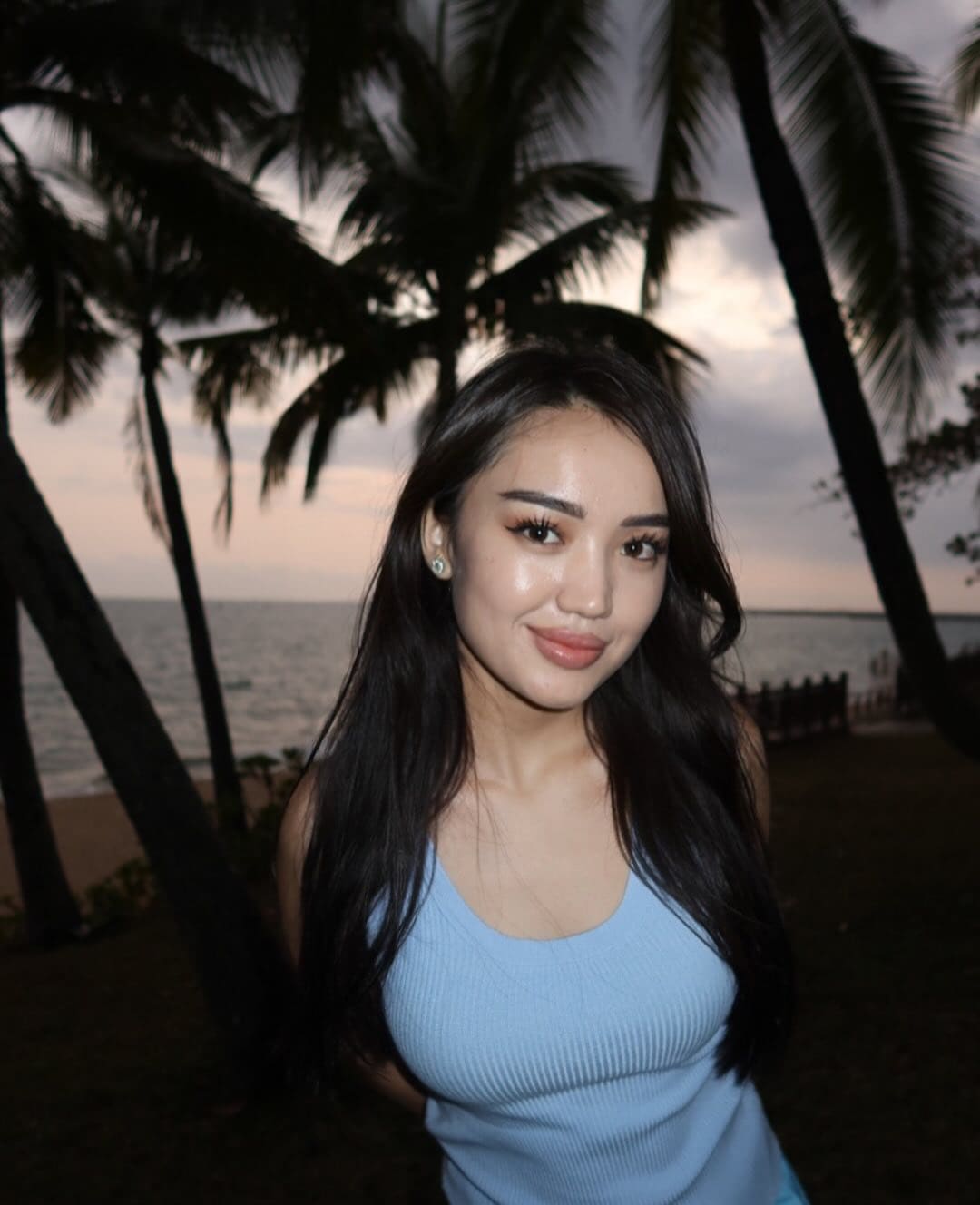 A woman with long dark hair stands outdoors at dusk near palm trees and the sea, wearing a light blue sleeveless ribbed top.
