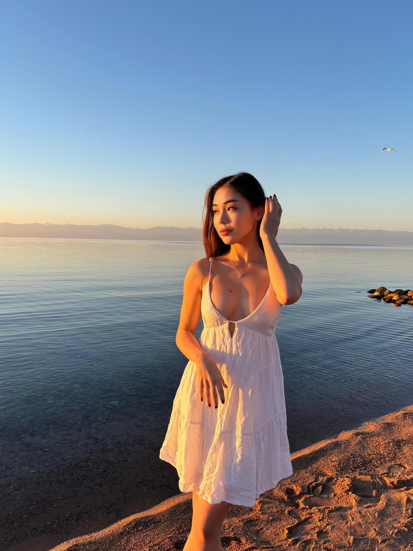 A woman stands on a sandy shore at sunset in a short white dress, one hand in her dark hair and the other resting on her thigh.