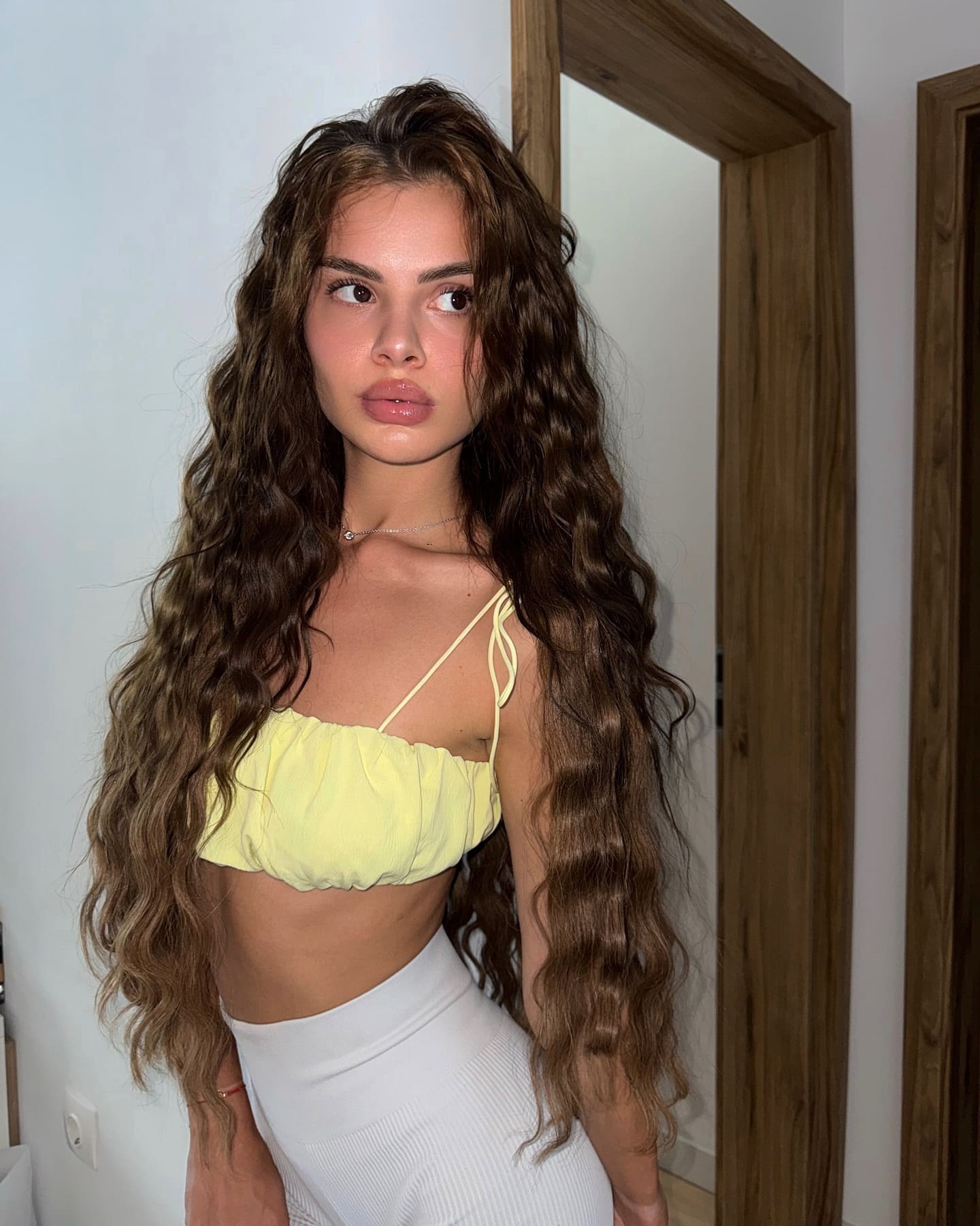 Iren stands indoors by a wooden door frame in a yellow strappy crop top and white high-waisted bottoms, head turned to the side.