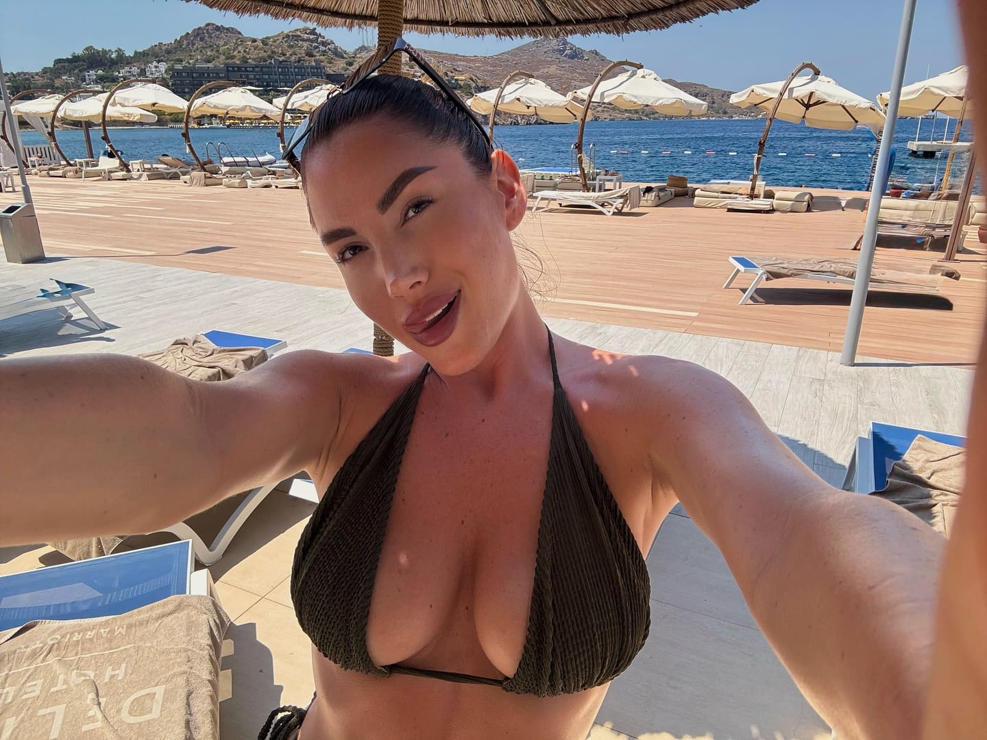 A woman takes a selfie in a dark green ribbed bikini top on a wooden beach deck, with white umbrellas and the sea behind her.