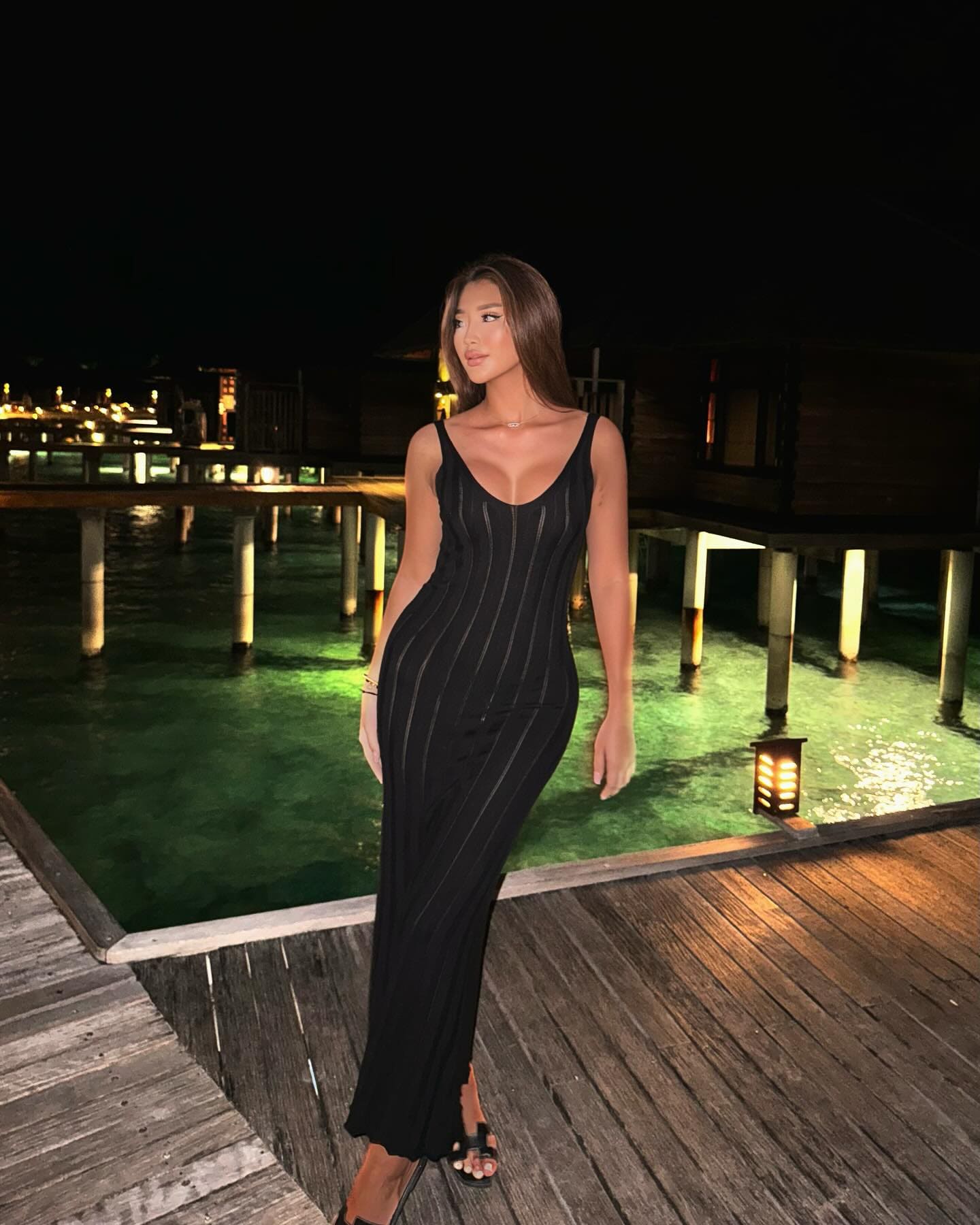 A woman in a black striped dress stands on a deck at night near illuminated water.