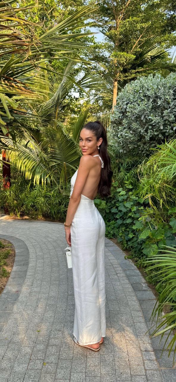 A woman in a white backless halter maxi dress stands on a paved garden path holding a white clutch, with tropical foliage in the background.