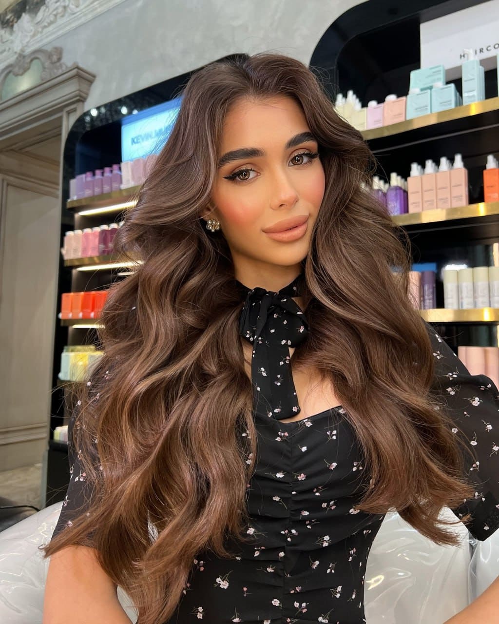 Ella sits in a salon chair wearing a black floral tie-neck dress, with shelves of hair products behind her.