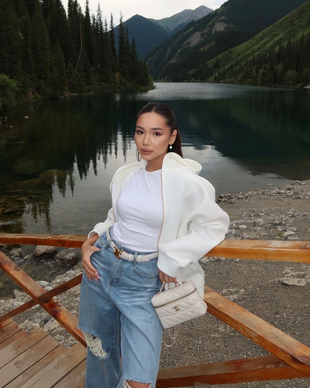 Woman in white t-shirt, jacket, and ripped jeans stands on a wooden deck by a mountain lake, holding a small quilted bag.