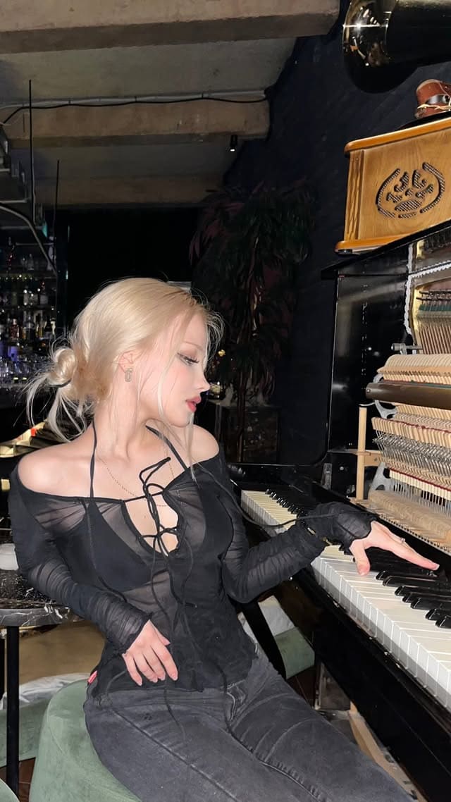 A woman with a low bun sits at a grand piano in a sheer black top and jeans, her hand on the keys in a dim room.